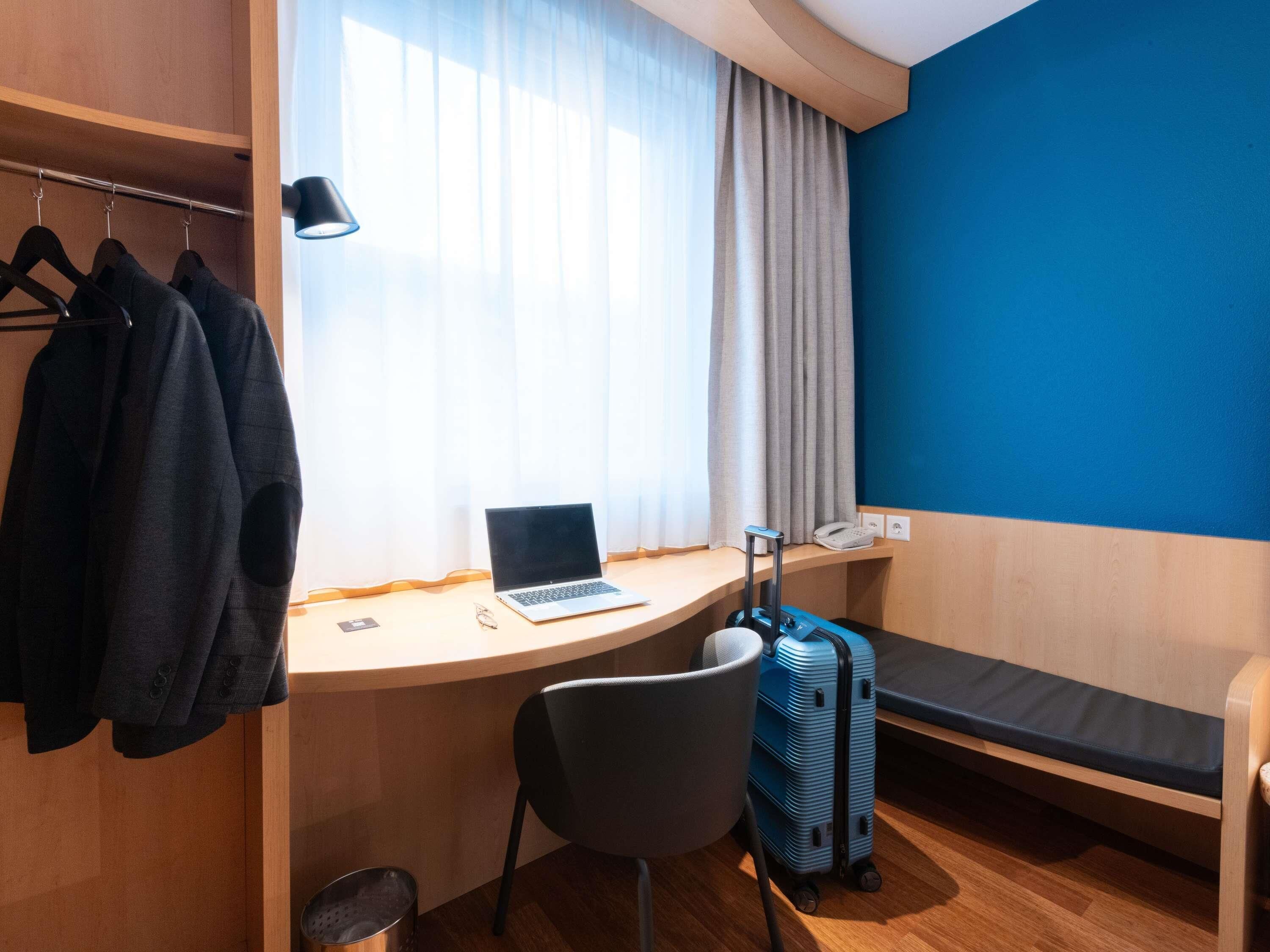 Habitación de hotel con escritorio, laptop, silla, cama, maleta y perchero con chaquetas.