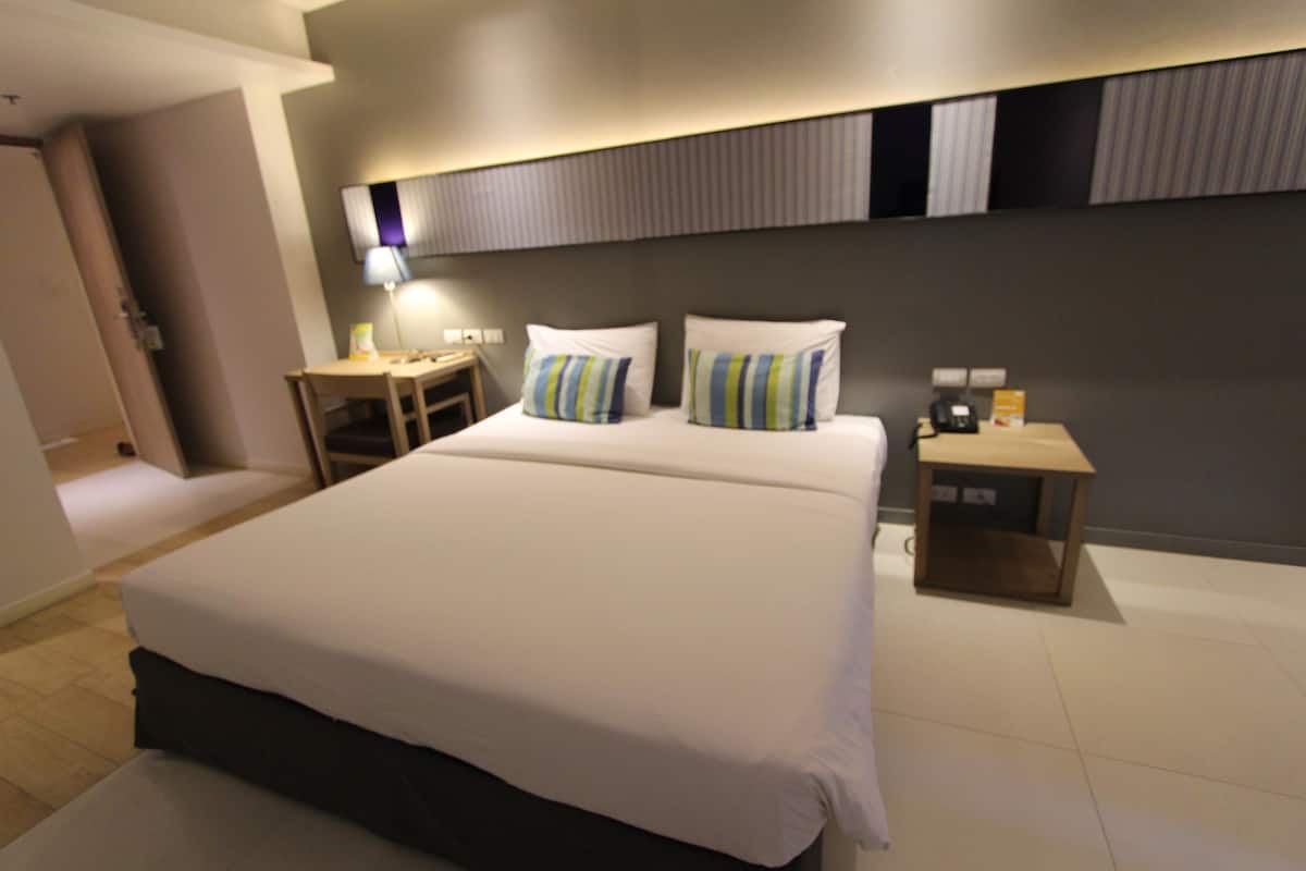 A modern hotel room with a large bed, two bedside tables, a nightstand, and a wall-mounted lamp.