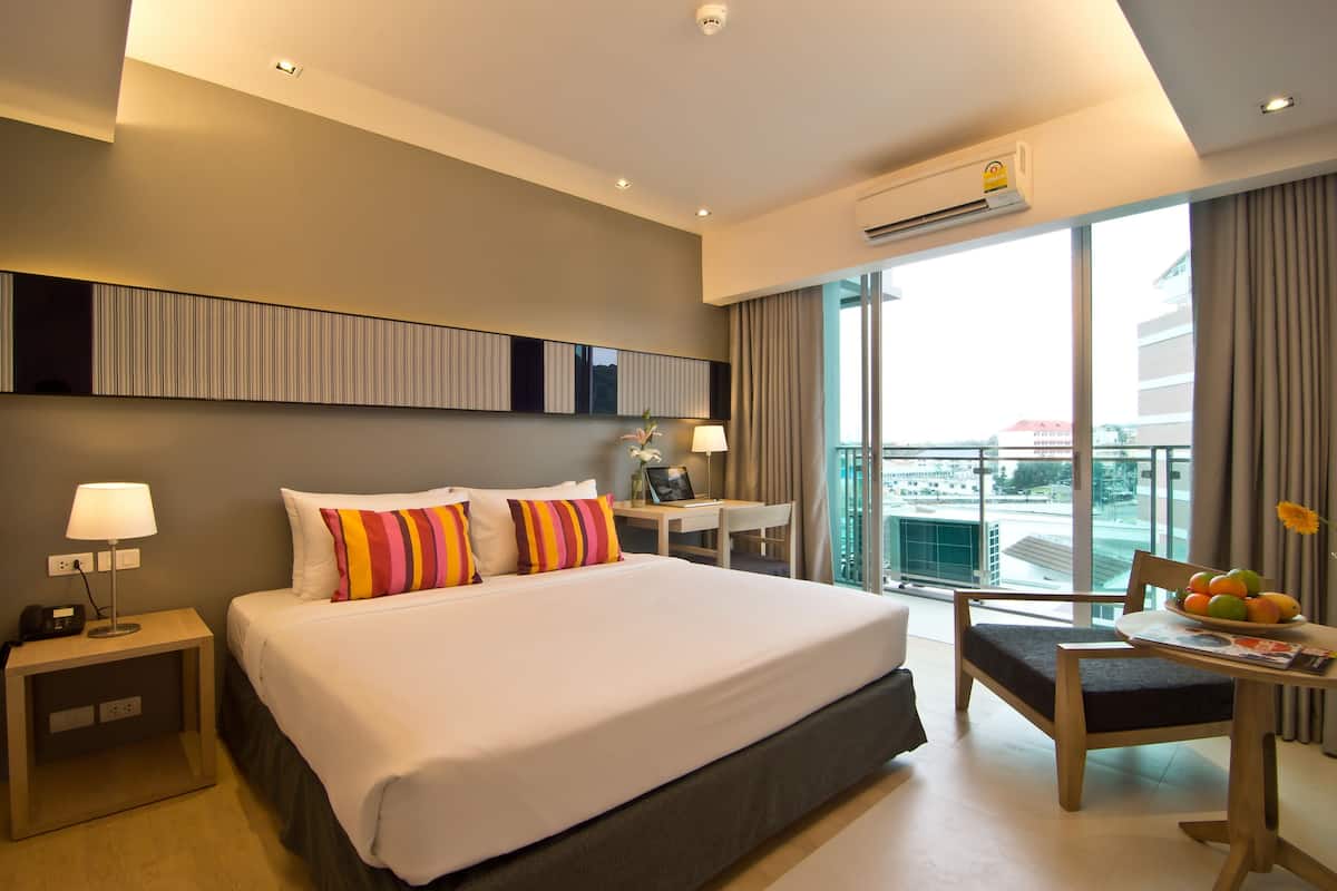 A hotel room with a large bed, a desk, a chair, and a view of the city through large windows.