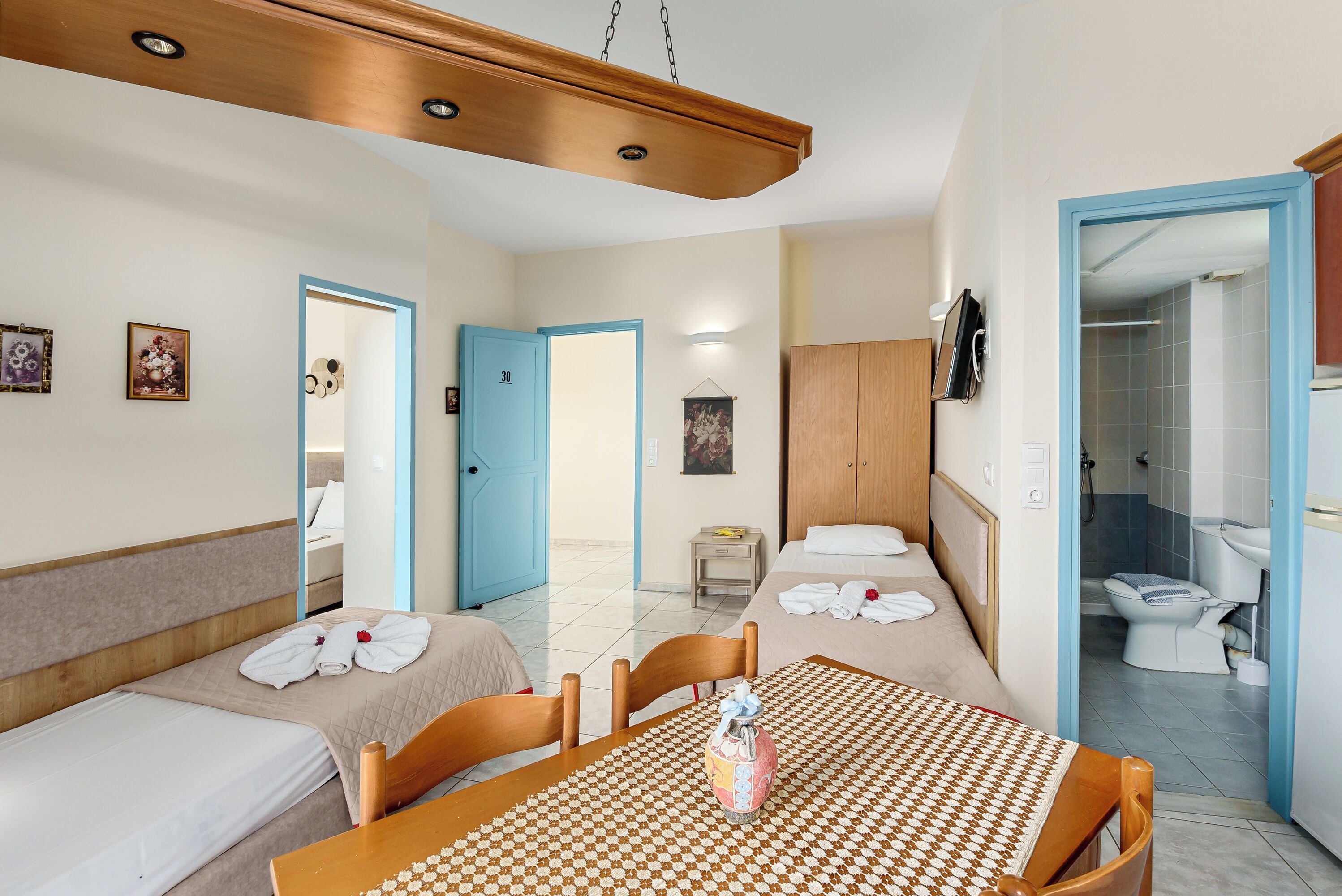 A hotel room with two beds, a wooden ceiling, a table with a checkered tablecloth, and a bathroom visible through an open door.
