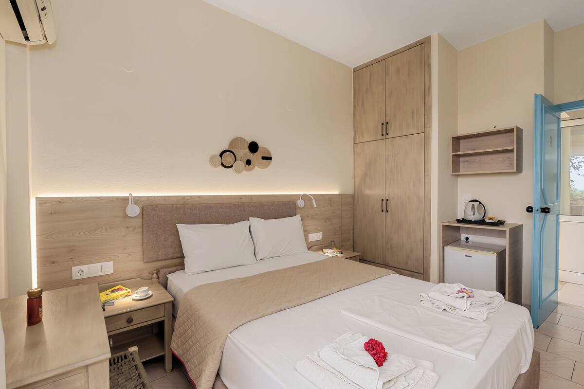 Une chambre d’hôtel avec un grand lit, une table de chevet avec une lampe, une commode, une armoire et un petit bureau avec une bouilloire.