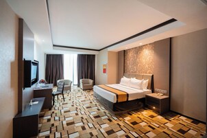 Quarto de hotel com uma cama, escrivaninha, cadeira, televisão e tapete estampado.