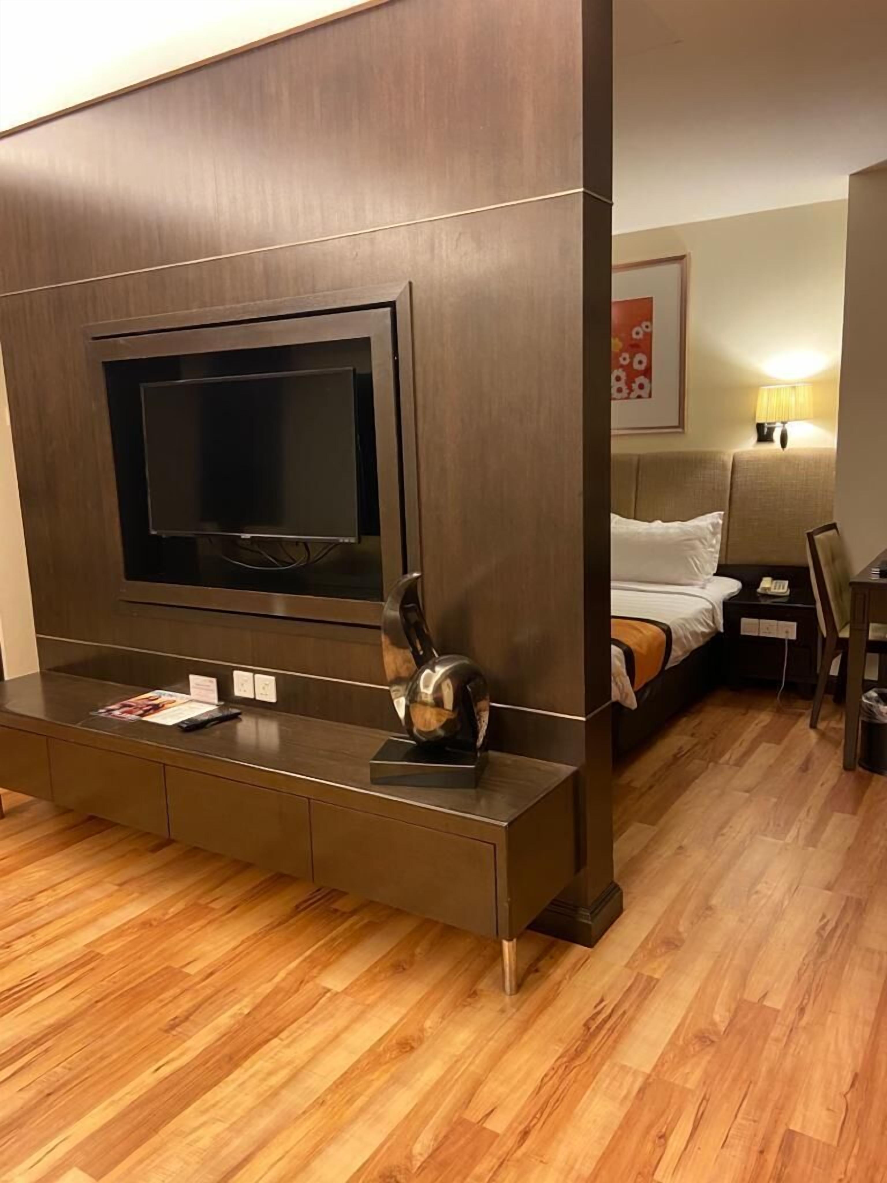 A modern hotel room with a large flat-screen TV mounted on a wooden panel, a wooden console table with a magazine, and a bed with a white comforter.