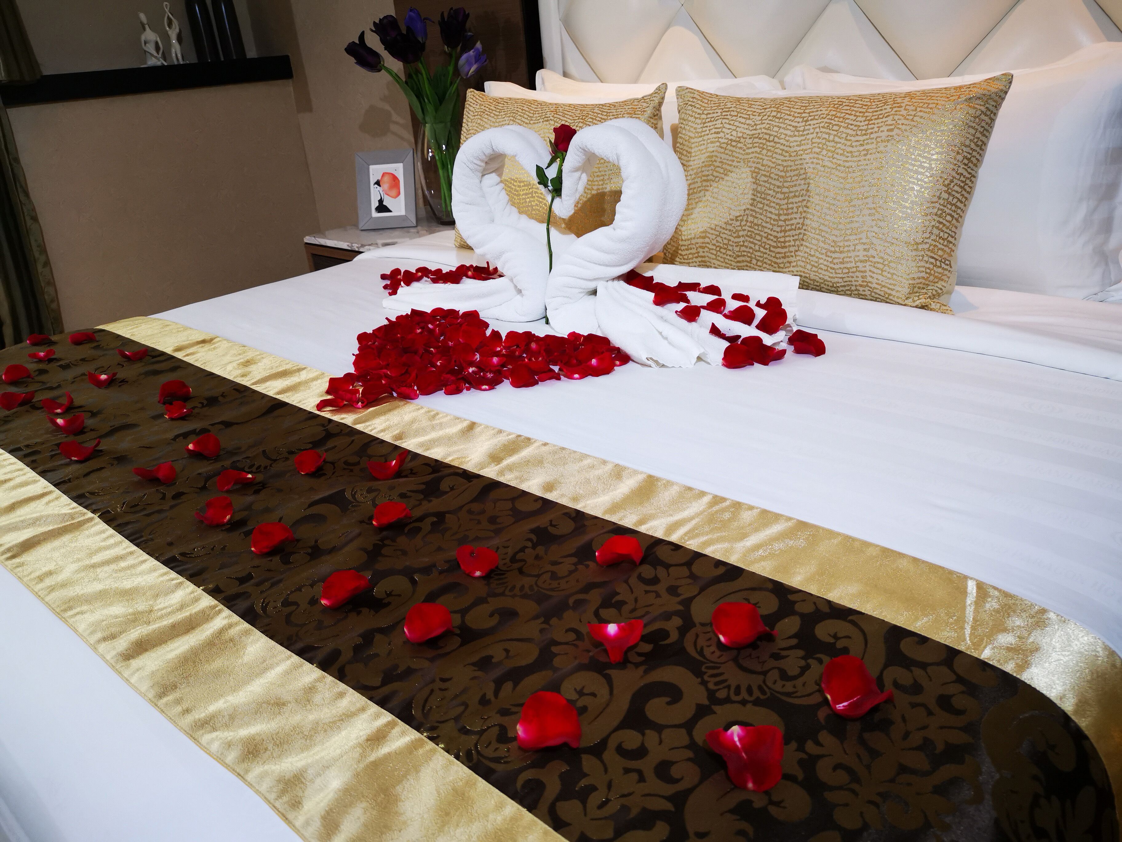 A bed with a white and gold bedspread, a bouquet of flowers, and a towel arranged in the shape of a swan.