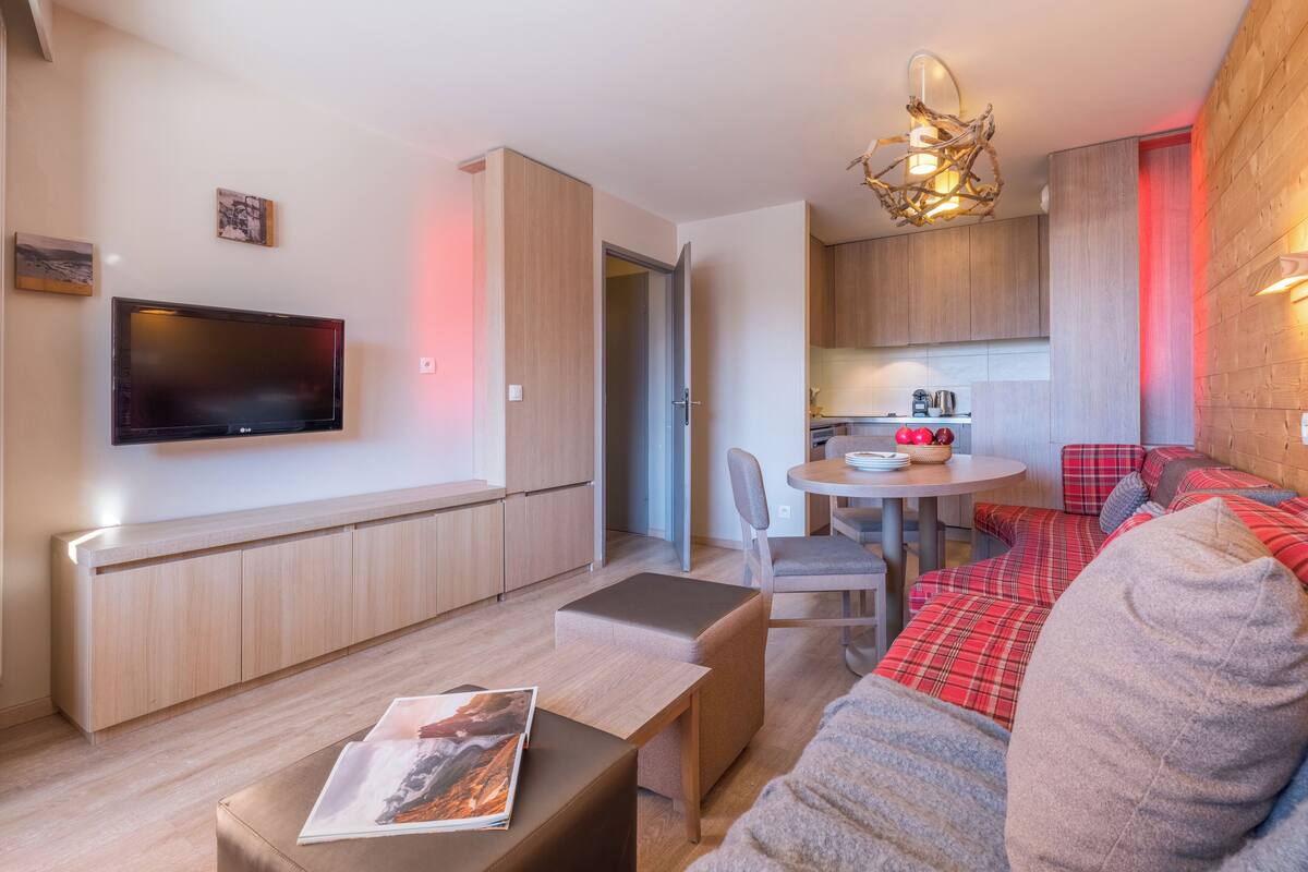 Una sala de estar moderna con un televisor de pantalla plana, una zona de comedor, una cocina y un rincón de descanso con un sofá rojo y gris.