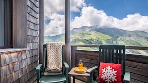 A balcony with wooden railings, two green chairs, a small table with a glass bottle and orange plate, and a red pillow with a snowflake design. - Résidence Pierre & Vacances Atria Crozats (Avoriaz)