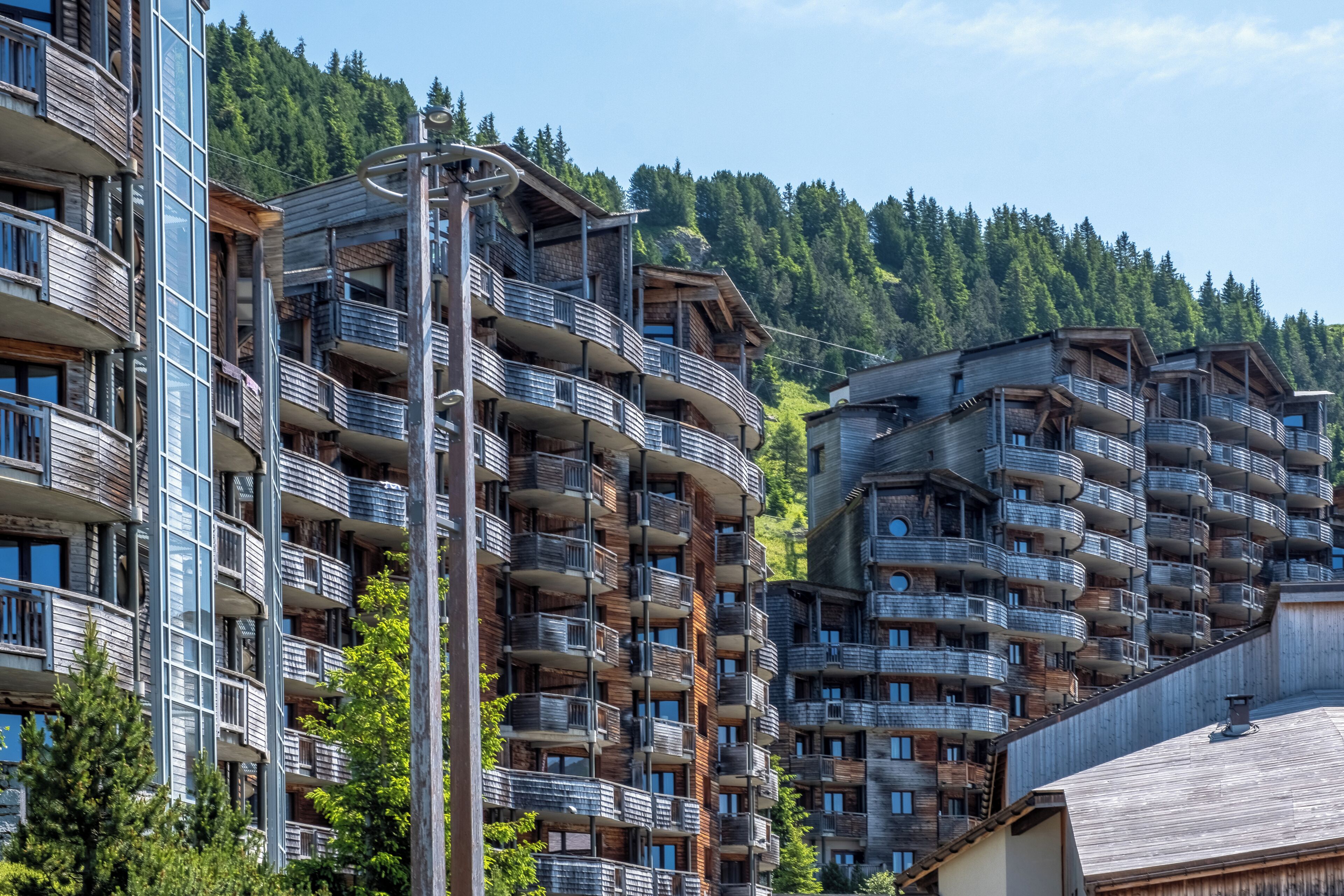 Un immeuble à plusieurs étages doté de balcons, d’une façade en bois et surplombant une colline boisée en arrière-plan.