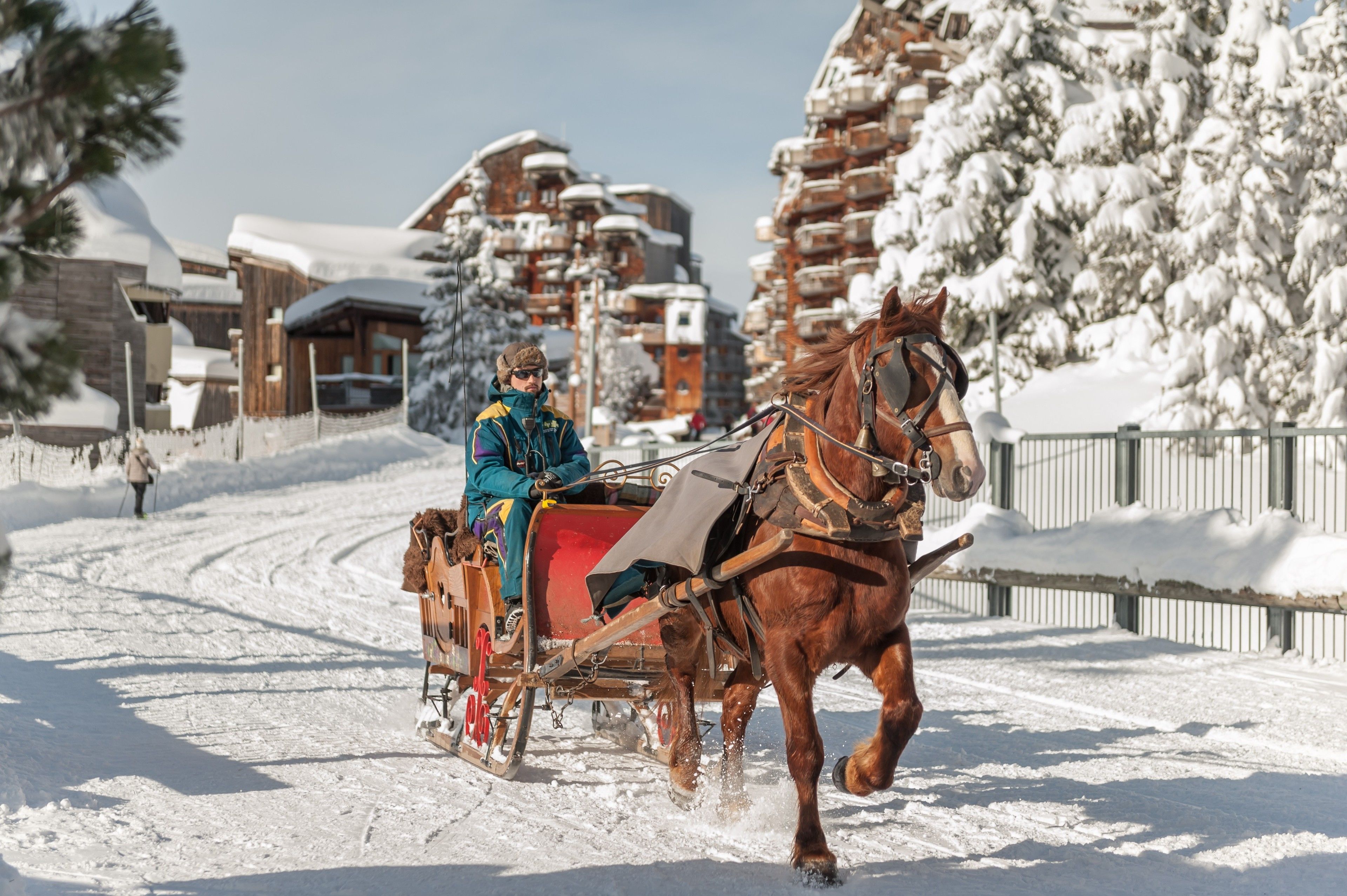 A horse-drawn sleigh on a snow-covered path with wooden buildings and snow-covered trees in the background.