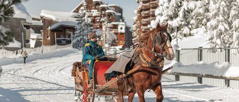A horse-drawn sleigh on a snow-covered path with wooden buildings and snow-covered trees in the background.