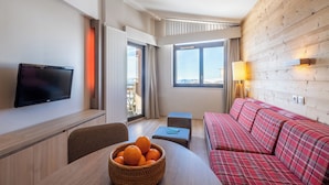 A modern hotel room with a flat-screen TV, a dining table with a basket of oranges, a red plaid sofa, and a large window offering a view. - Résidence Pierre & Vacances Atria Crozats (Avoriaz)