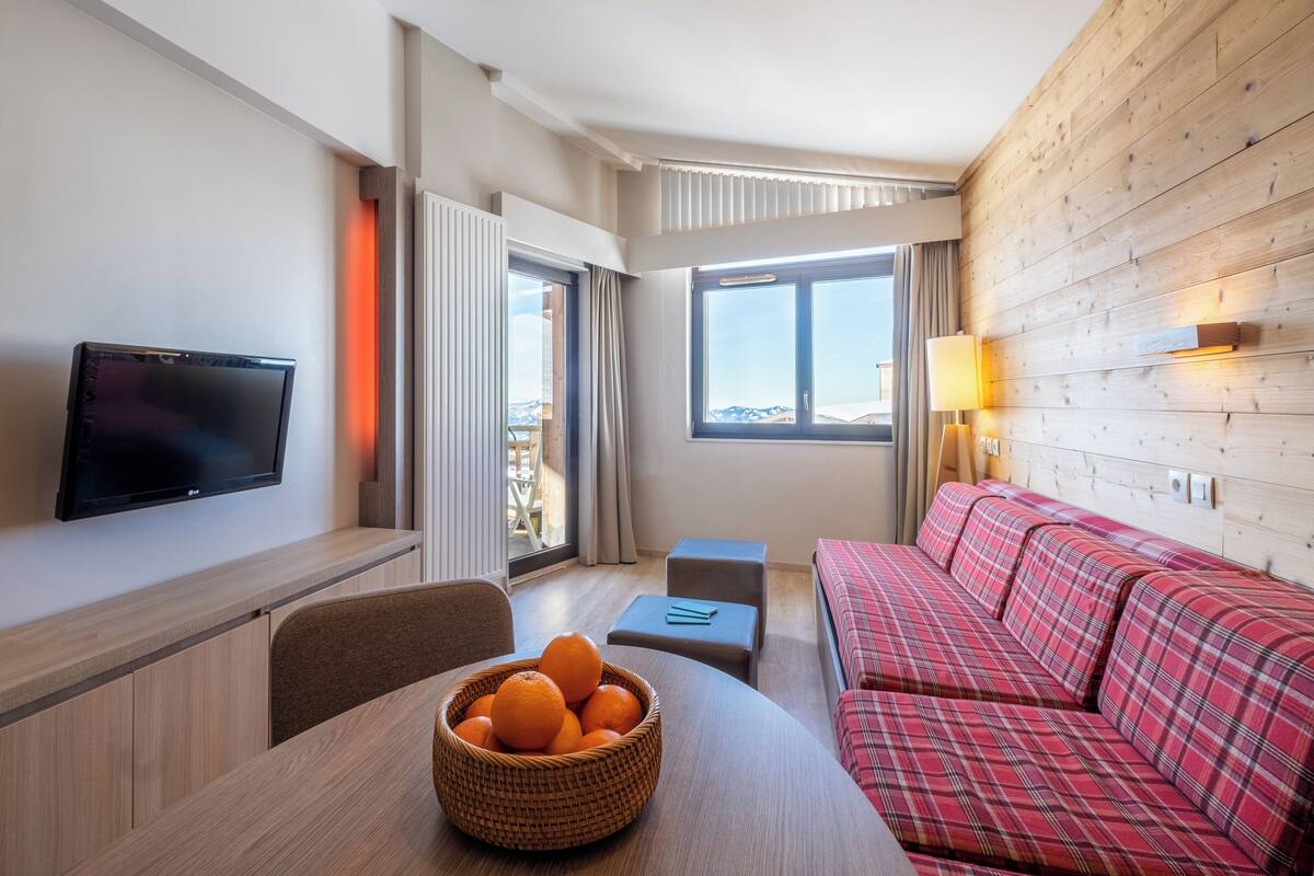 Habitación de hotel moderna con una televisión de pantalla plana, una mesa de comedor con una cesta de naranjas, un sofá rojo a cuadros y un ventanal con vistas.