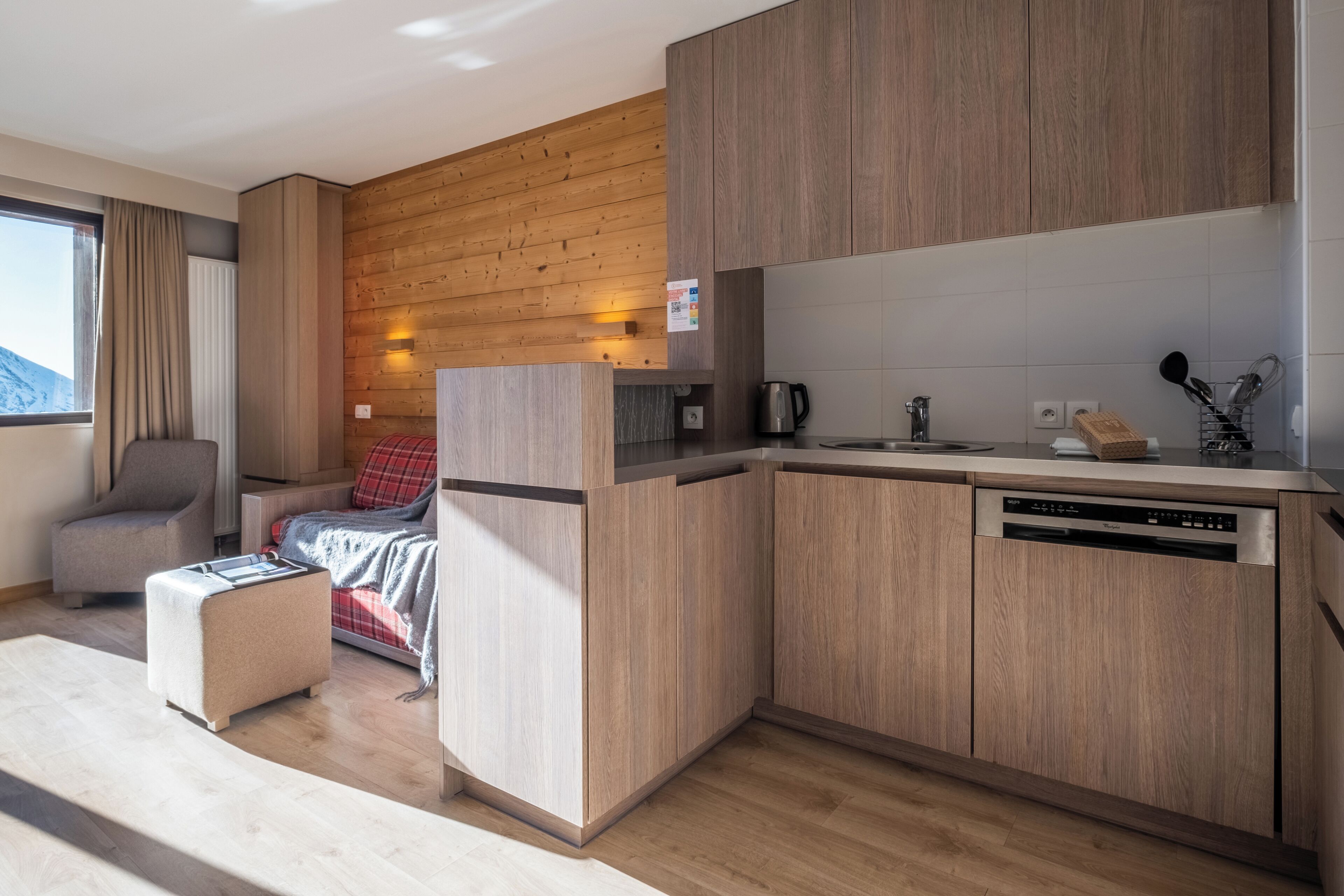 Ein kleiner Raum mit Küchenzeile, einem Bett mit roter Decke, einem grauen Sessel und einer Holzwand.