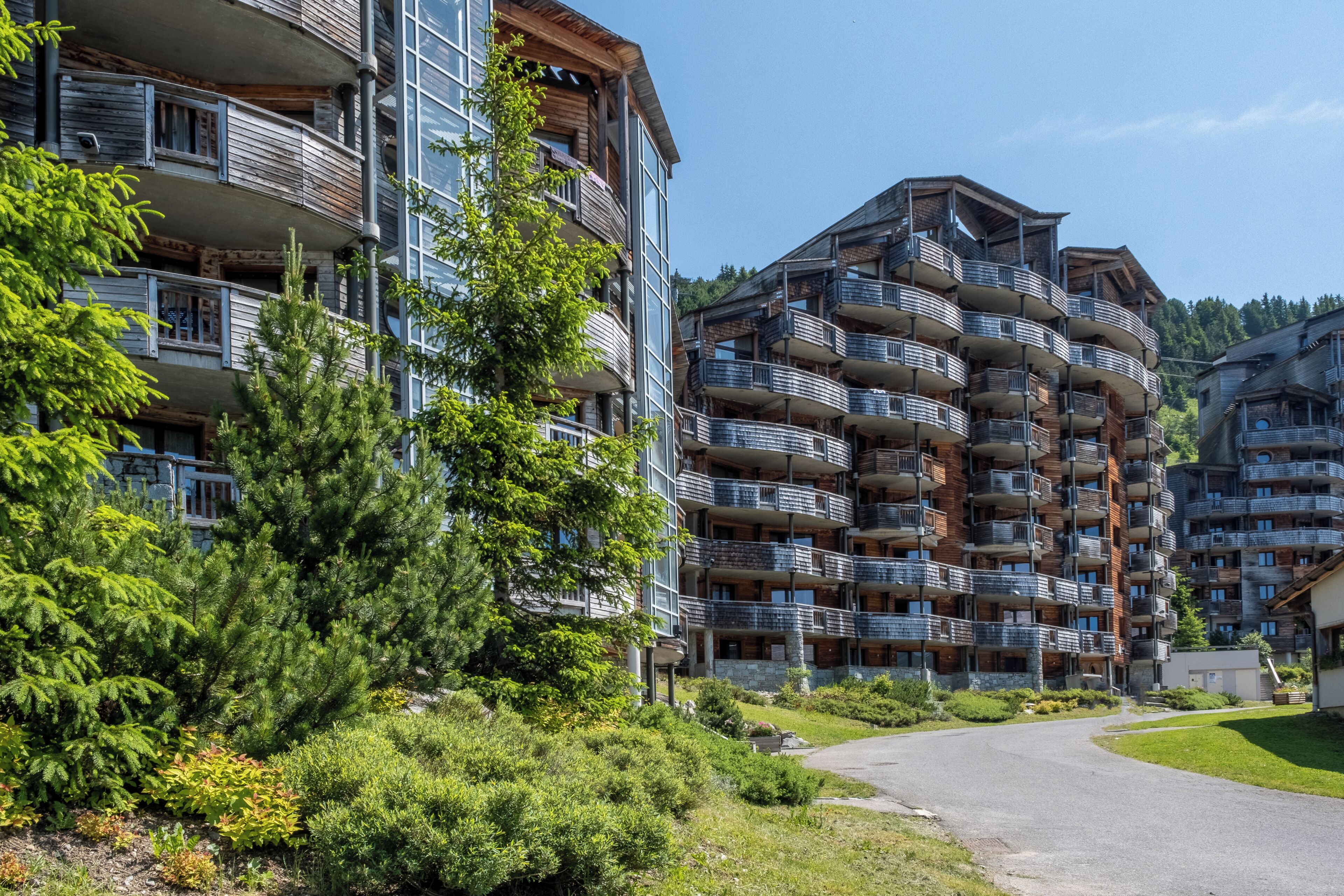 A multi-story building with balconies, surrounded by greenery and a clear sky.