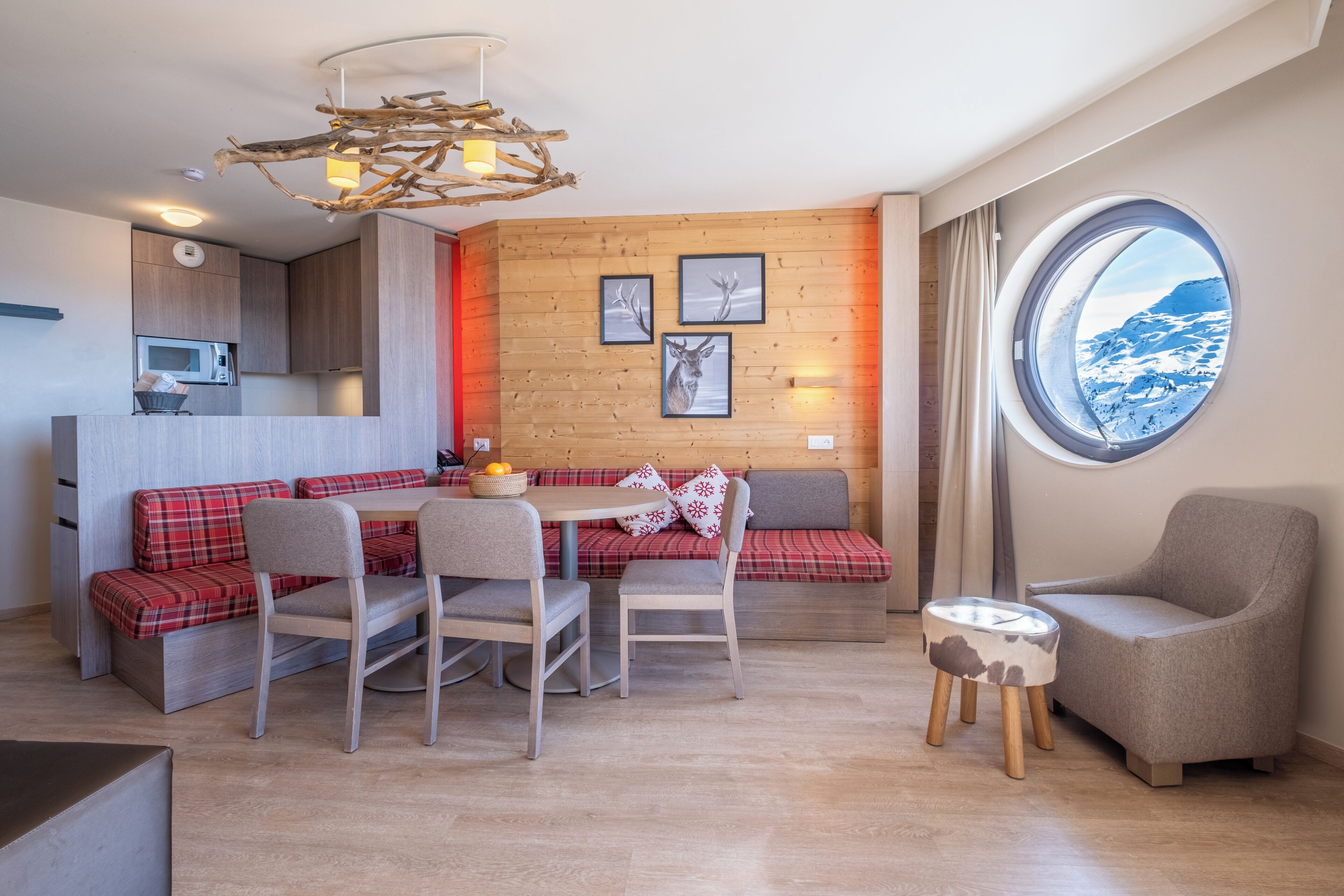 Une cuisine moderne dotée d’un four à micro-ondes encastré, un coin repas avec une table et des chaises, un banc à carreaux rouges et une fenêtre ronde offrant une vue sur les montagnes.