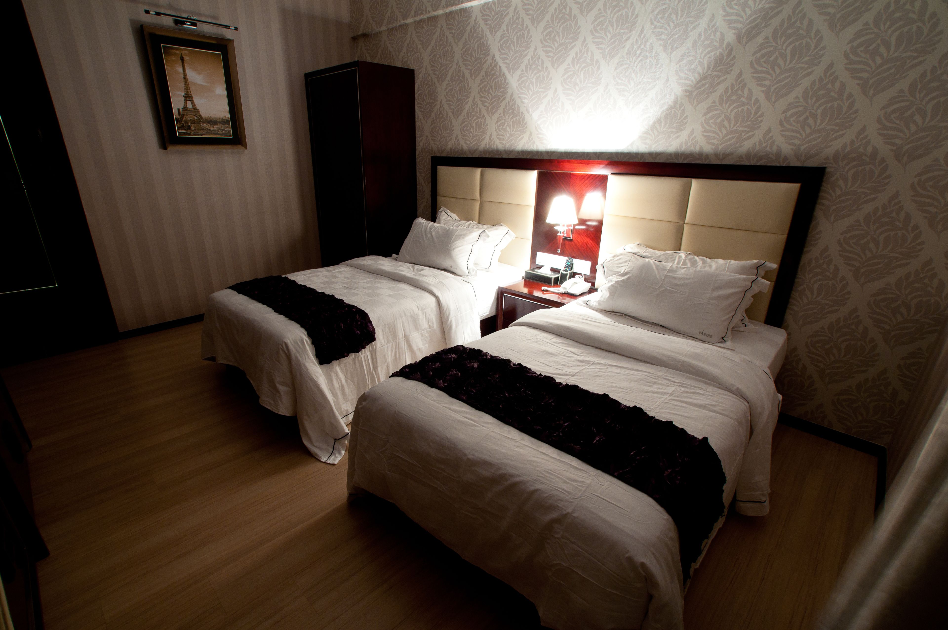 Habitación de hotel con dos camas, una mesita de noche con lámpara, un armario y un cuadro enmarcado en la pared.