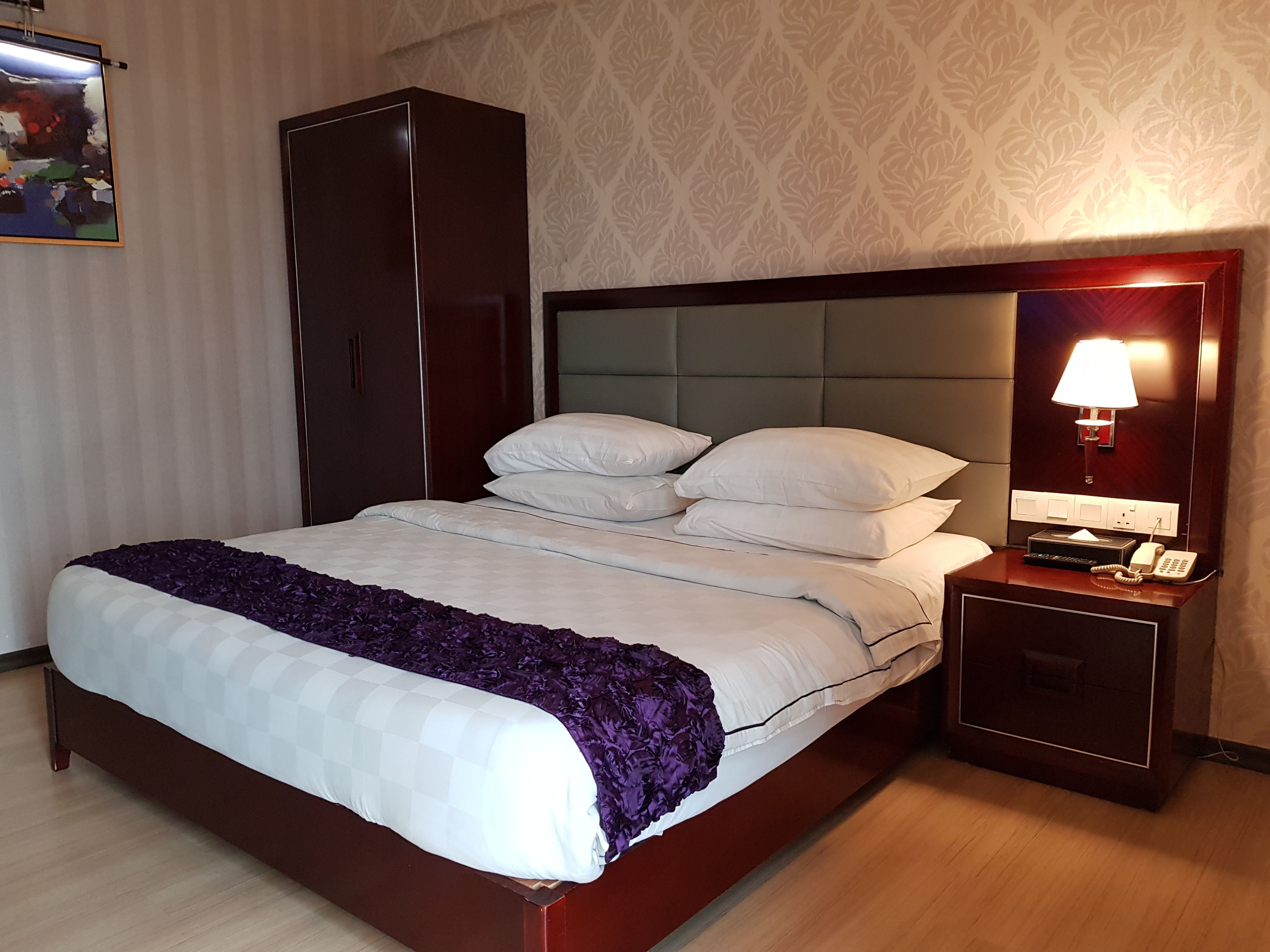 A neatly made bed with white and purple bedding, a wooden headboard, a nightstand with a lamp, and a telephone.