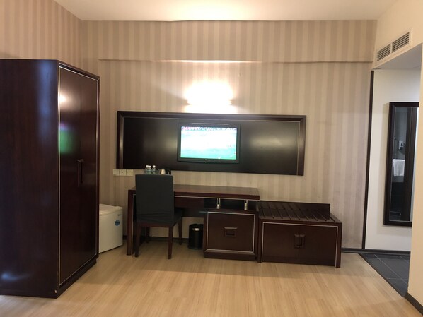 A hotel room with a large flat-screen TV mounted on the wall, a wooden desk with drawers, a chair, and a wardrobe.