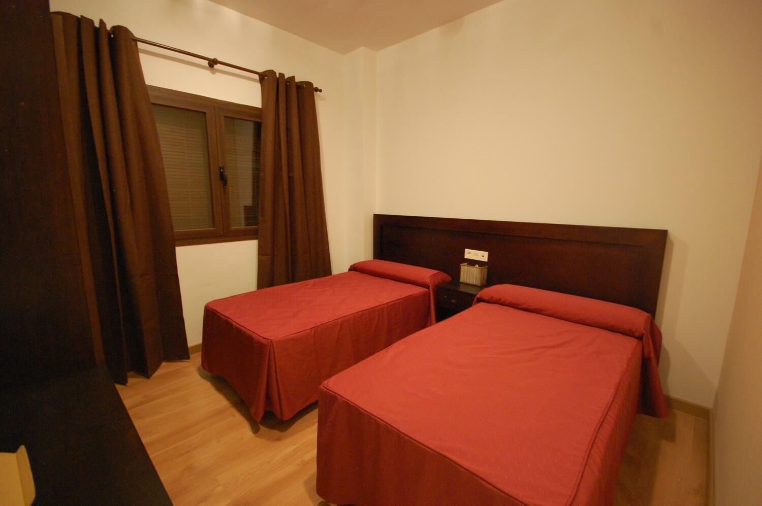 Una habitación con dos camas individuales, una ventana con cortinas, una mesita de noche con lámpara y suelo de madera.