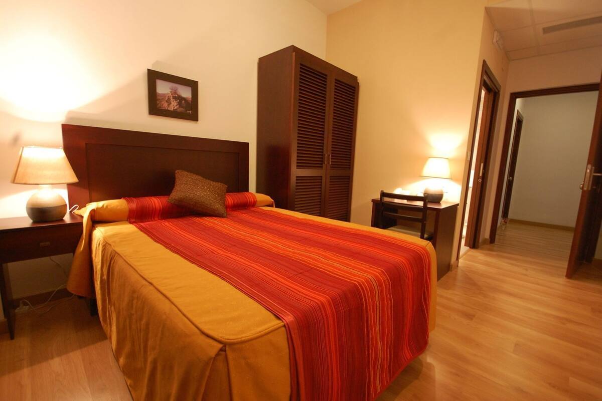 A hotel room with a bed, two bedside tables with lamps, a wooden headboard, a wardrobe, and a door leading to another room.