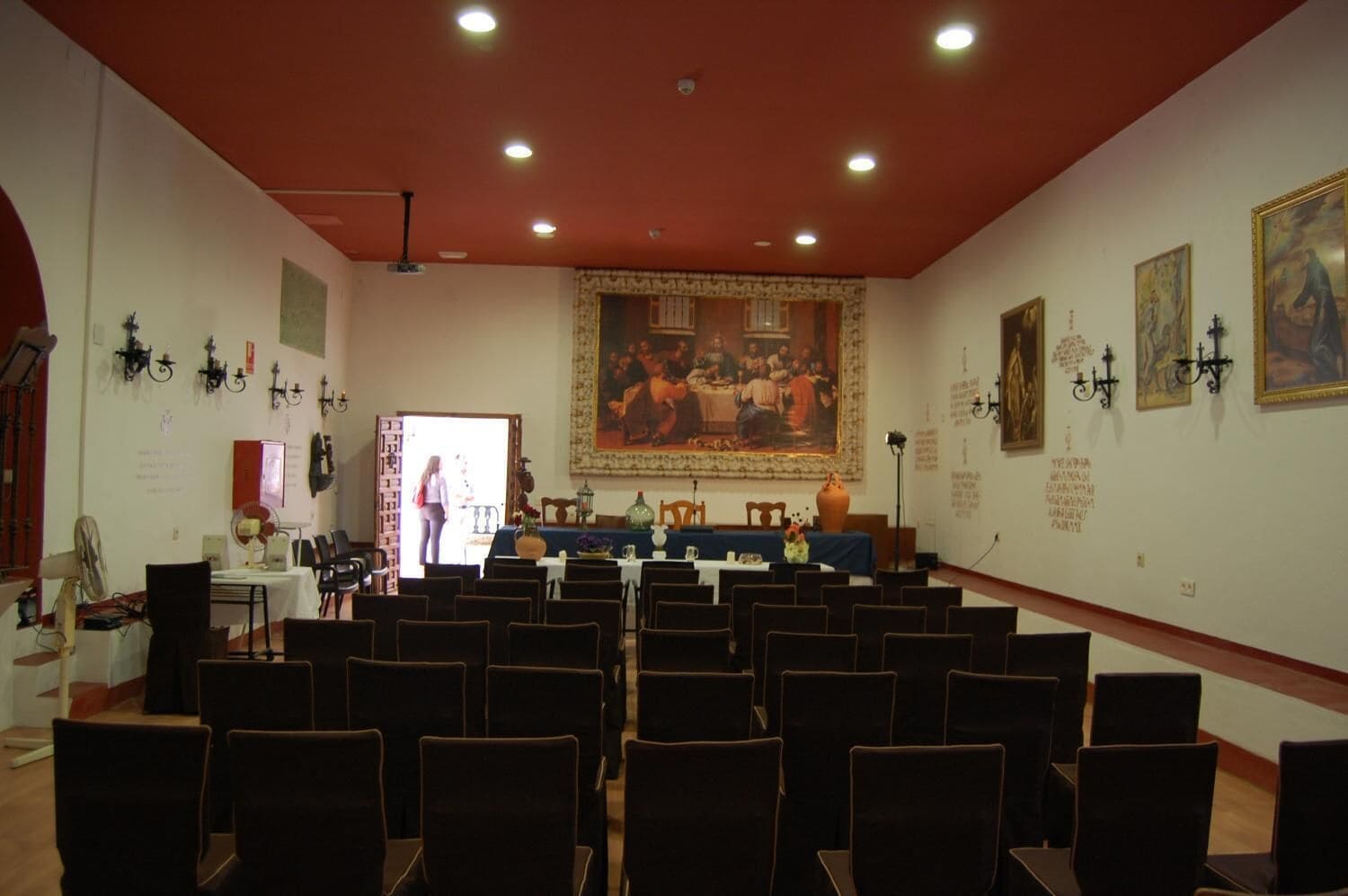 Un salón de conferencias con filas de sillas orientadas hacia un escenario, un cuadro grande en la pared y luminarias fijadas a la pared.