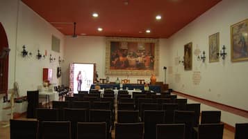 Un salón de conferencias con filas de sillas orientadas hacia un escenario, un cuadro grande en la pared y luminarias fijadas a la pared.