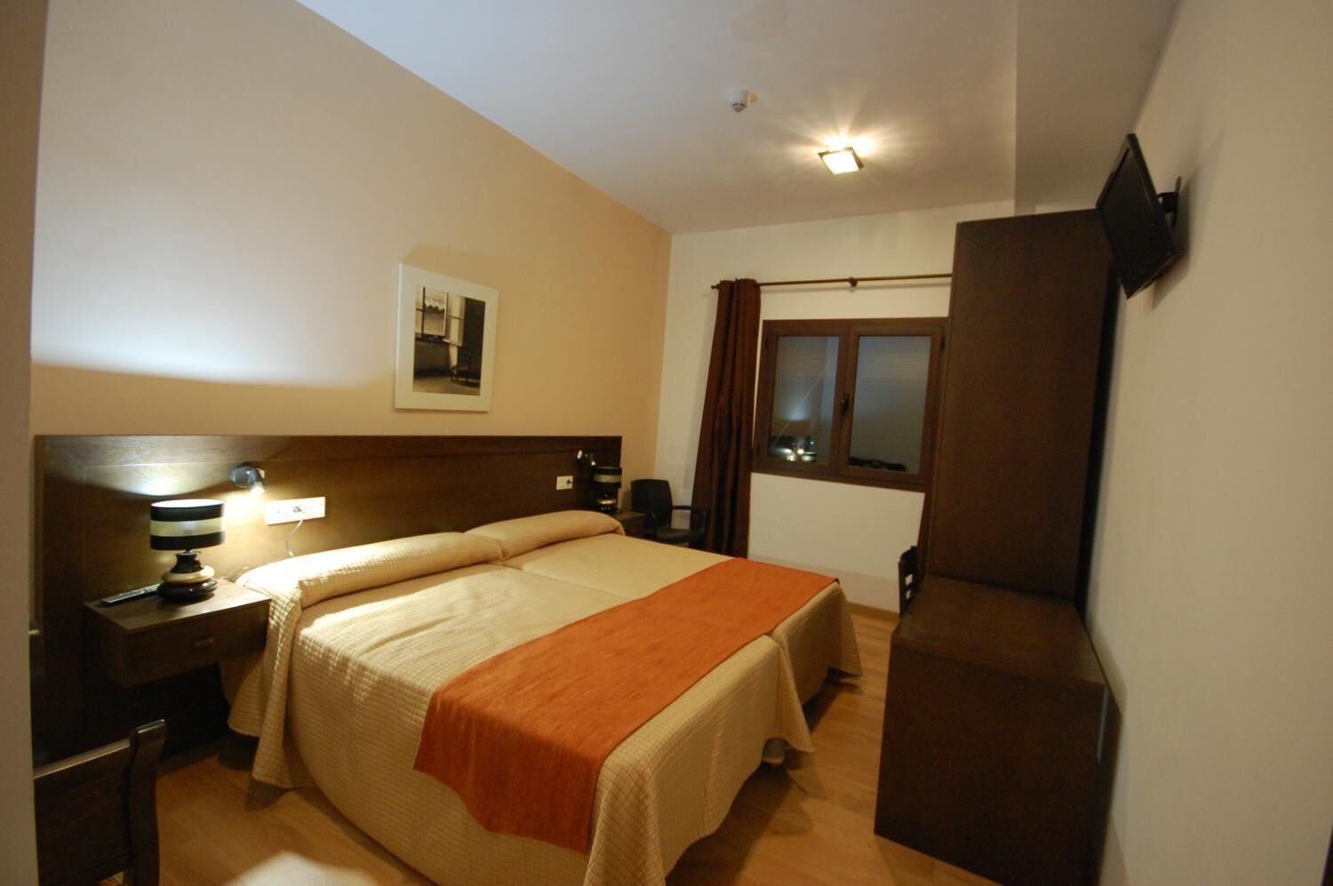Una habitación de hotel con cama, mesita de noche, lámpara y ventana con cortinas.