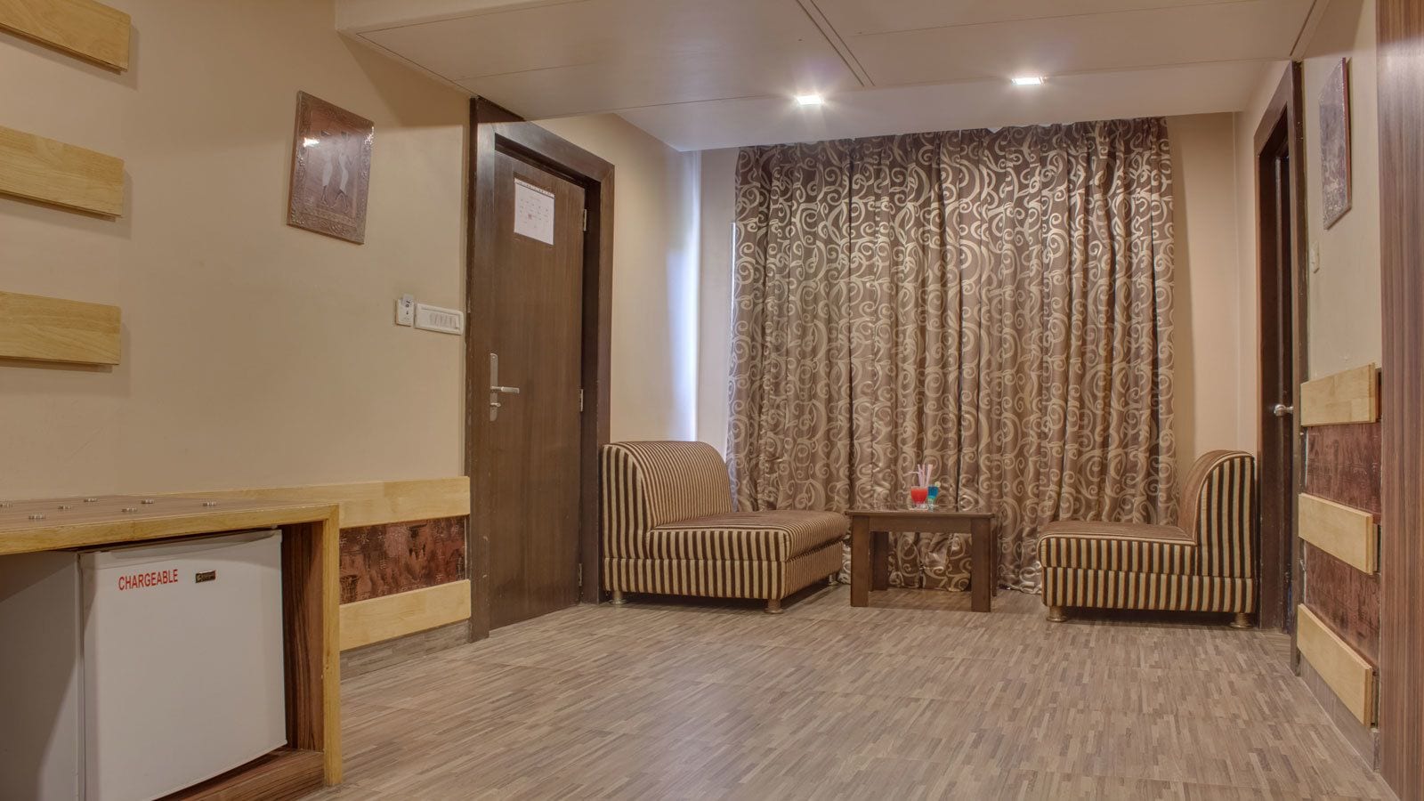 Una habitación con un escritorio de madera, dos sillones a rayas, una mesita con una bebida y una cortina estampada.