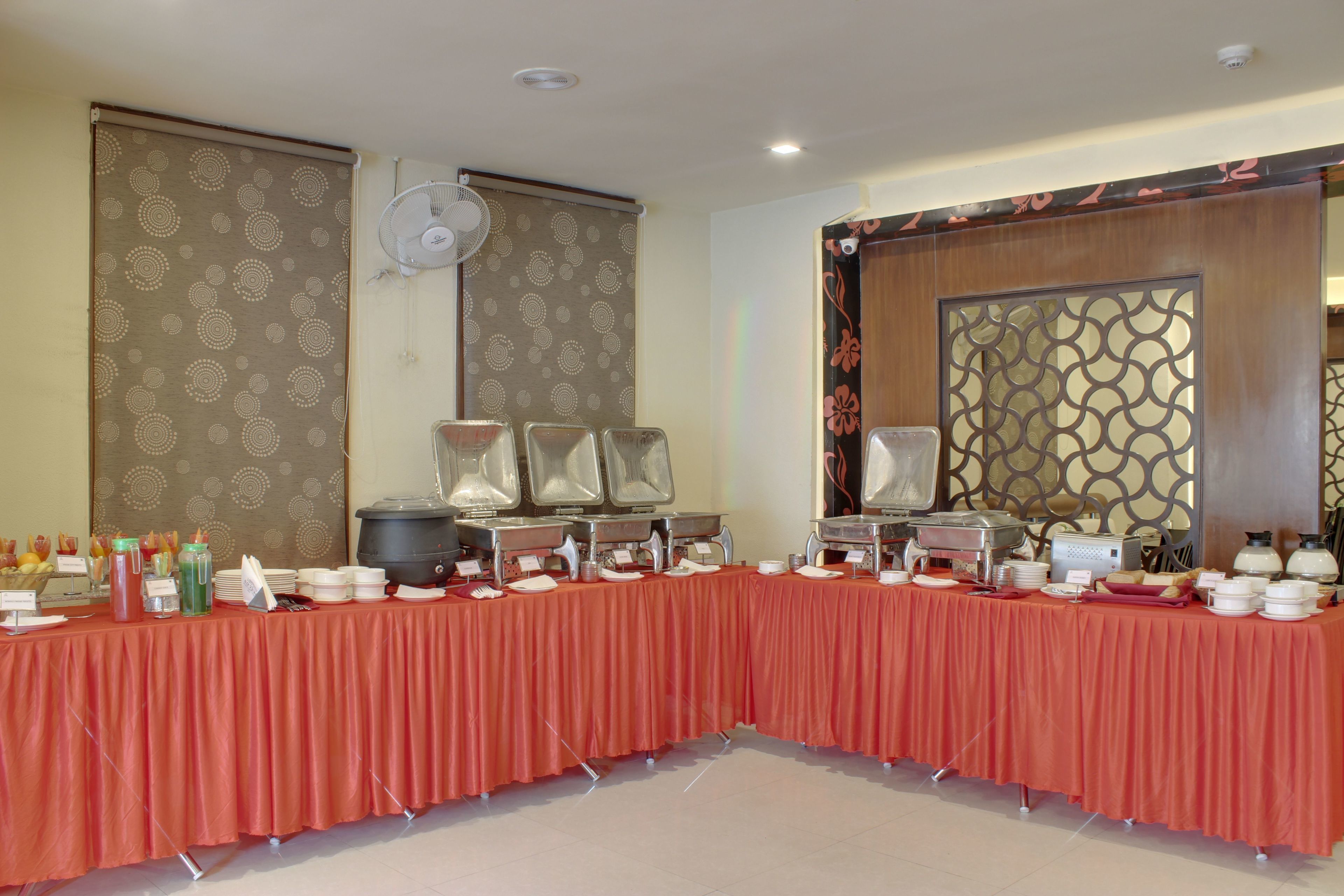 Montaje de bufé con una larga mesa cubierta con mantel rojo, equipada con diferentes recipientes de comida y utensilios para servir.