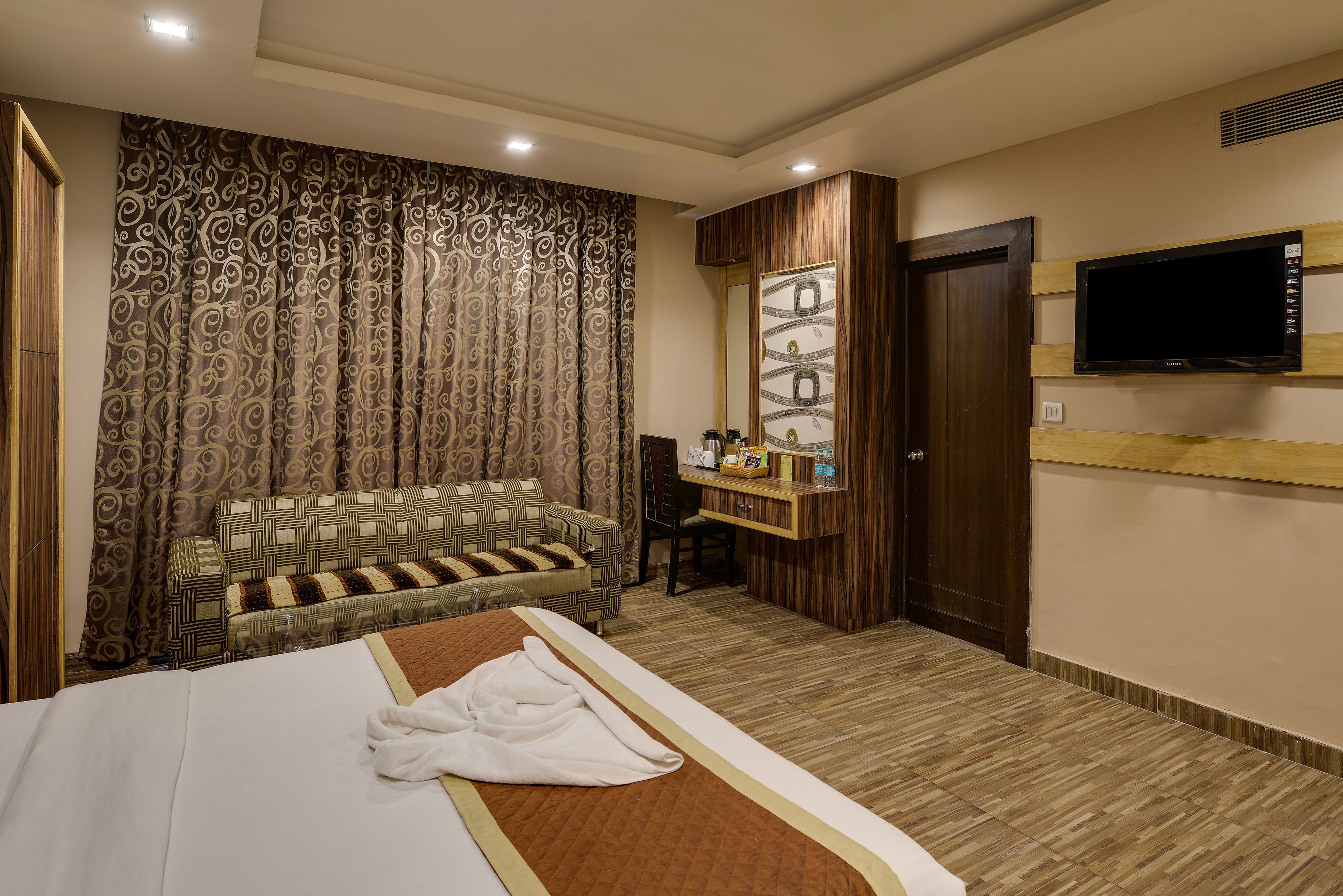 A hotel room with a bed, a sofa, a TV, and a small desk with a mirror and a shelf.