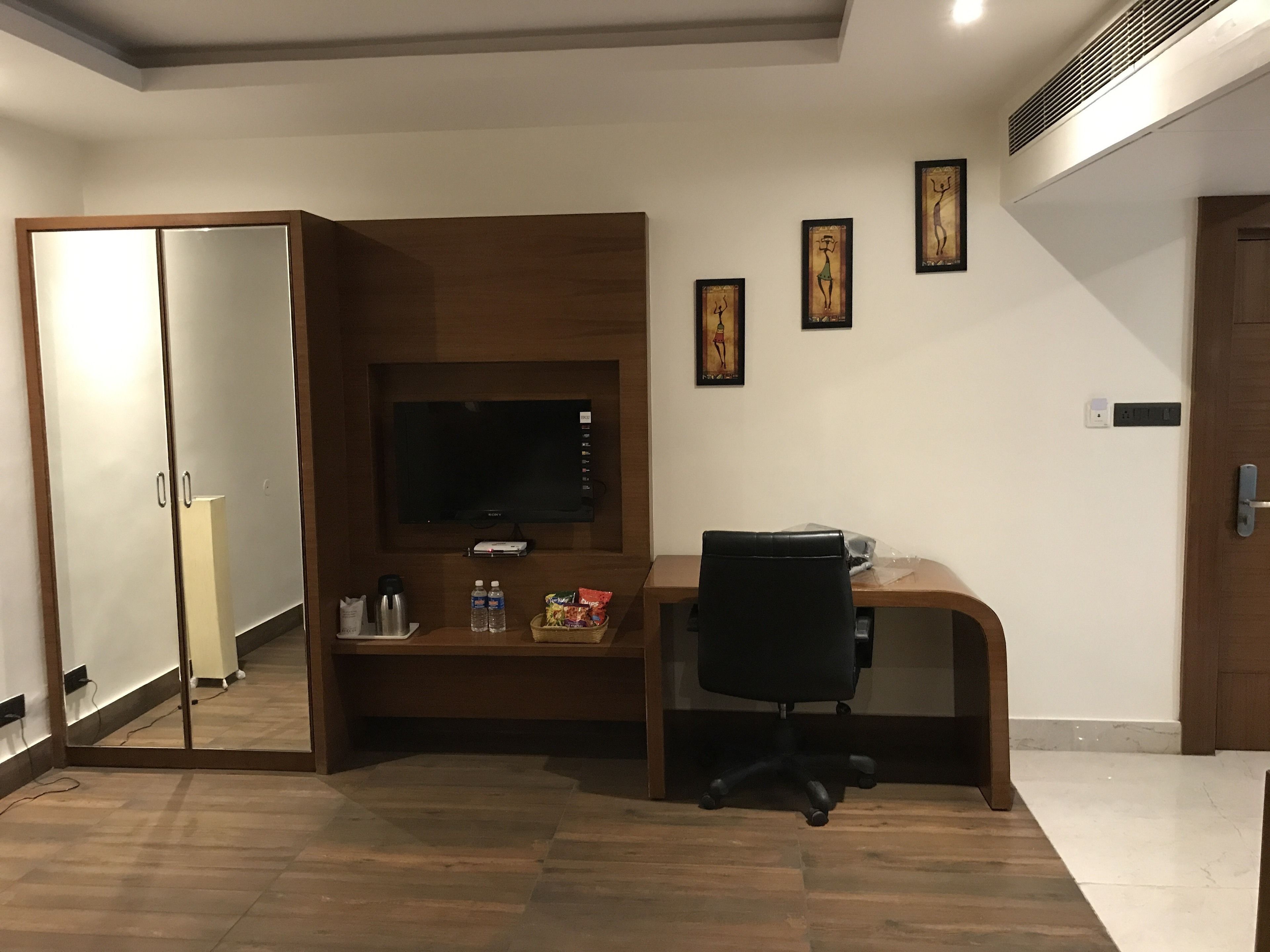 Un espacio de oficina moderno con un escritorio de madera, una silla de oficina negra, un televisor montado y una estantería pequeña con objetos personales.