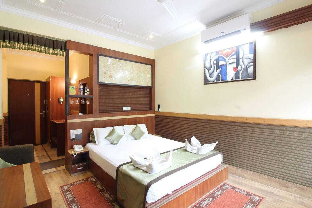 A hotel room with a bed, bedside table, a mirror, a shelf with toiletries, a painting on the wall, and a door leading to another room.