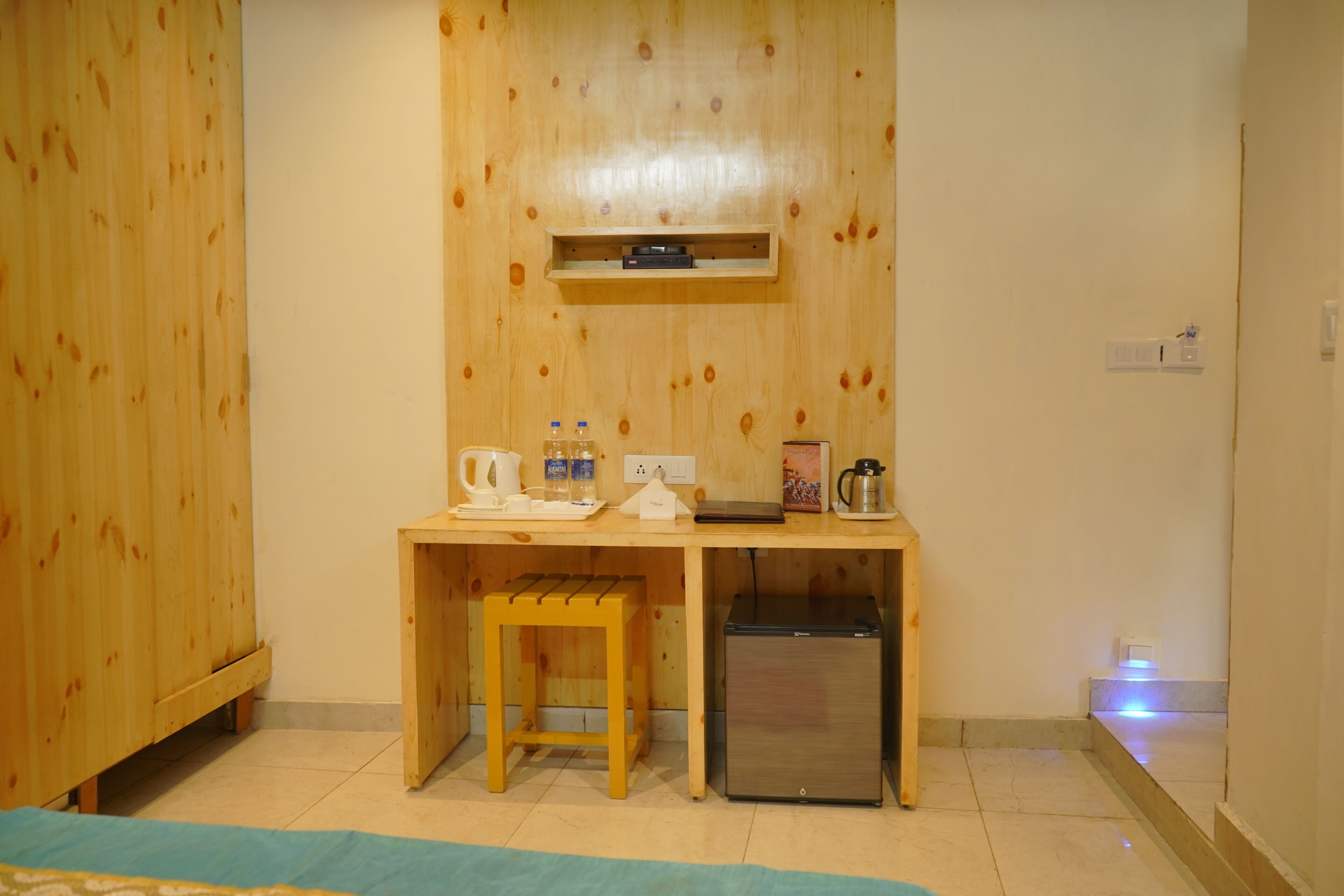Una habitación compacta con un escritorio de madera, un taburete pequeño, un mini refrigerador y una estantería sobre el escritorio.