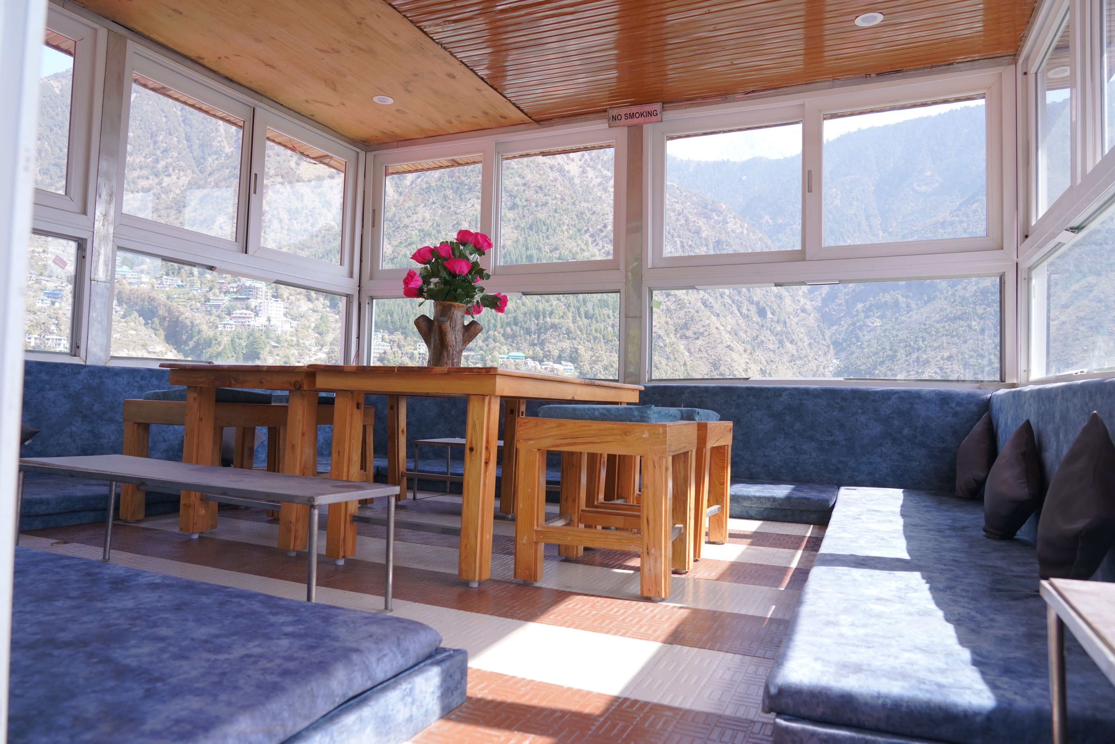 Una habitación con una mesa de madera, bancos y un sofá con cojines. Se ve una ventana con vistas a las montañas.