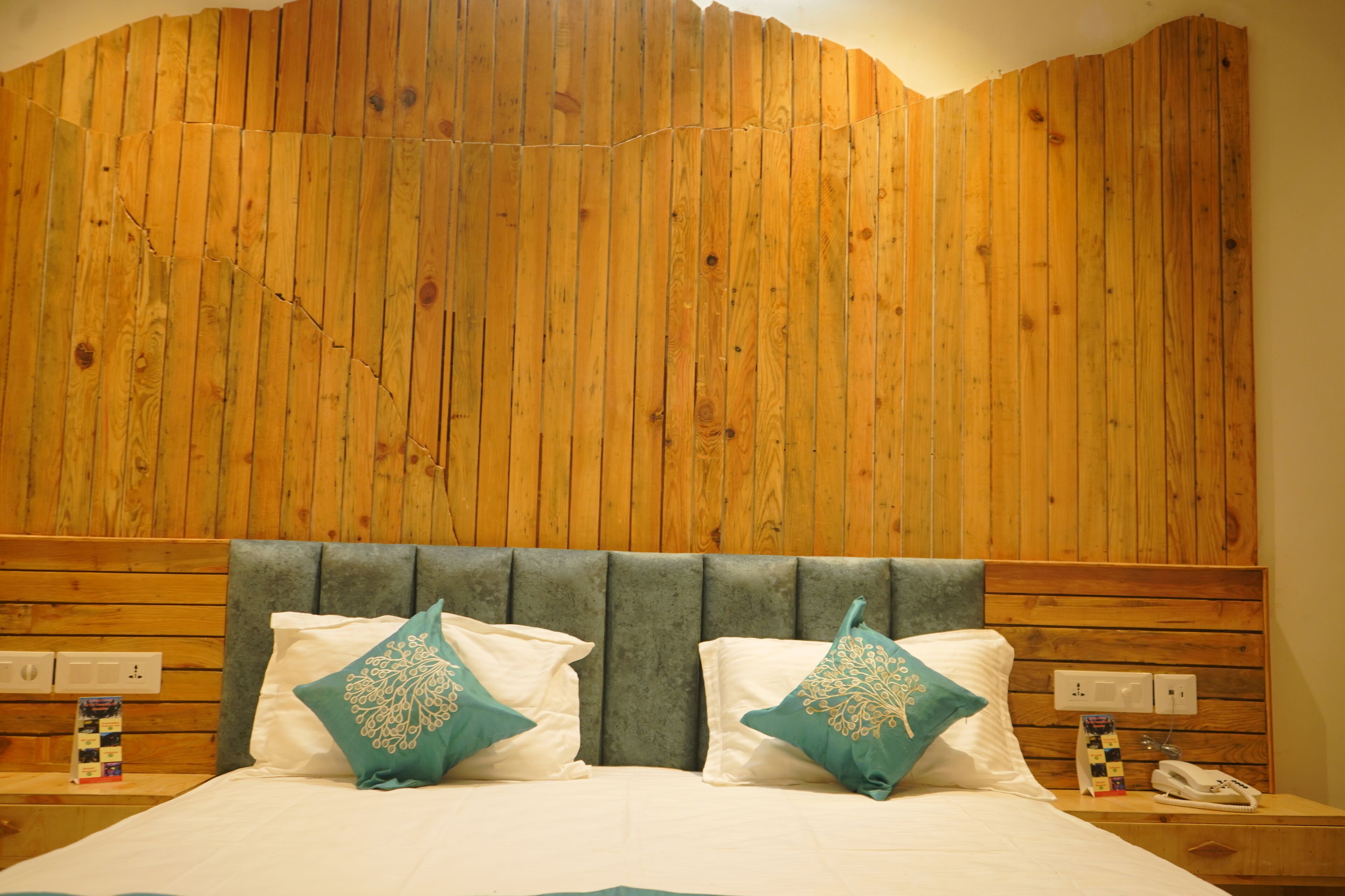 A hotel room with a wooden headboard, two pillows with a floral design, a bedside table with a phone, and a wall socket.
