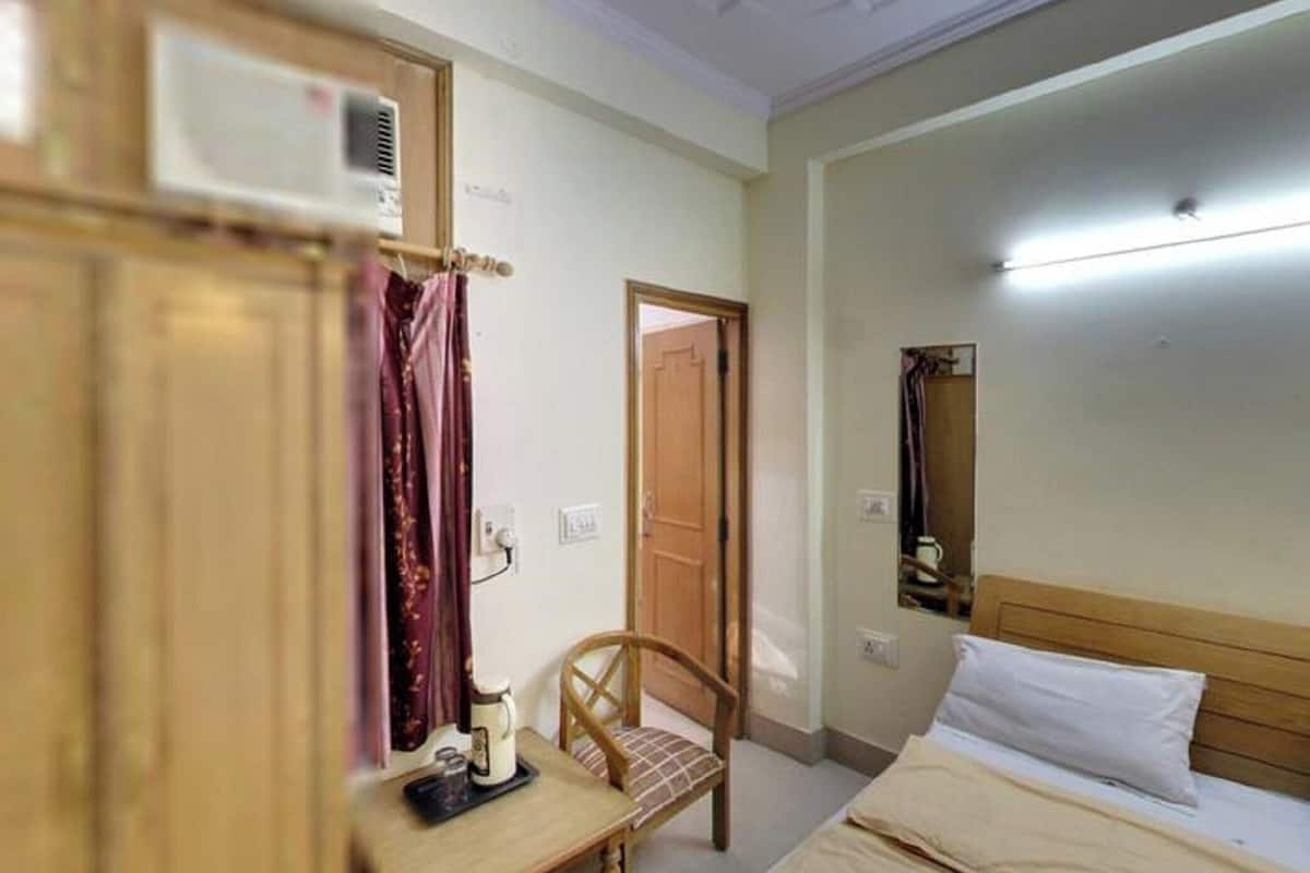 A hotel room with a bed, a chair, a desk with a lamp, a mirror, and an air conditioning unit.