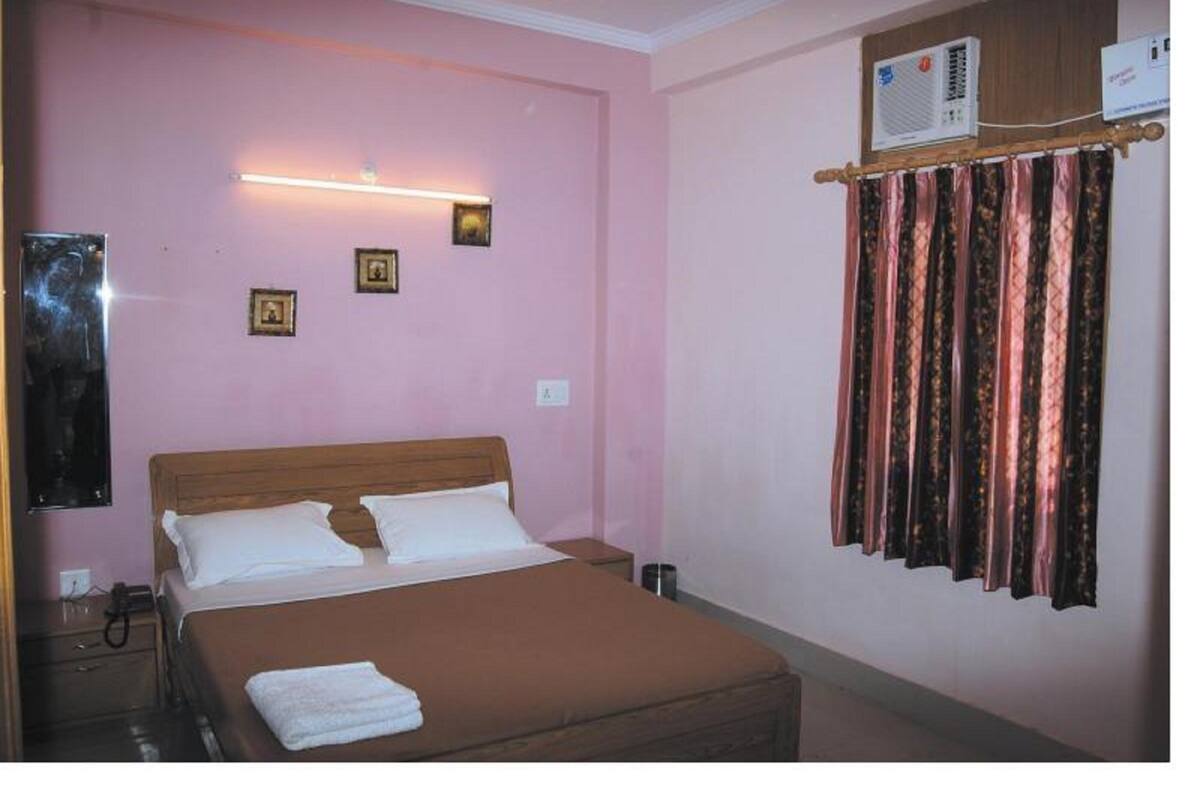 A bedroom with a bed, a bedside table, a wall-mounted air conditioner, and a window with curtains.