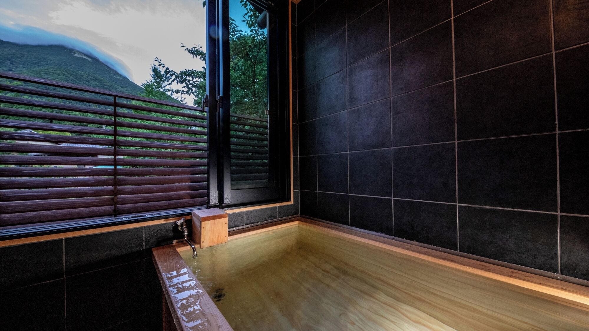 A modern bathroom with a wooden bench, a large window with blinds, and dark tiled walls.