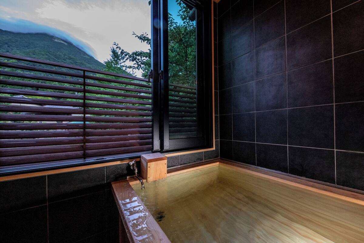 Un baño moderno con un banco de madera, un ventanal con persianas y paredes revestidas de baldosas oscuras.