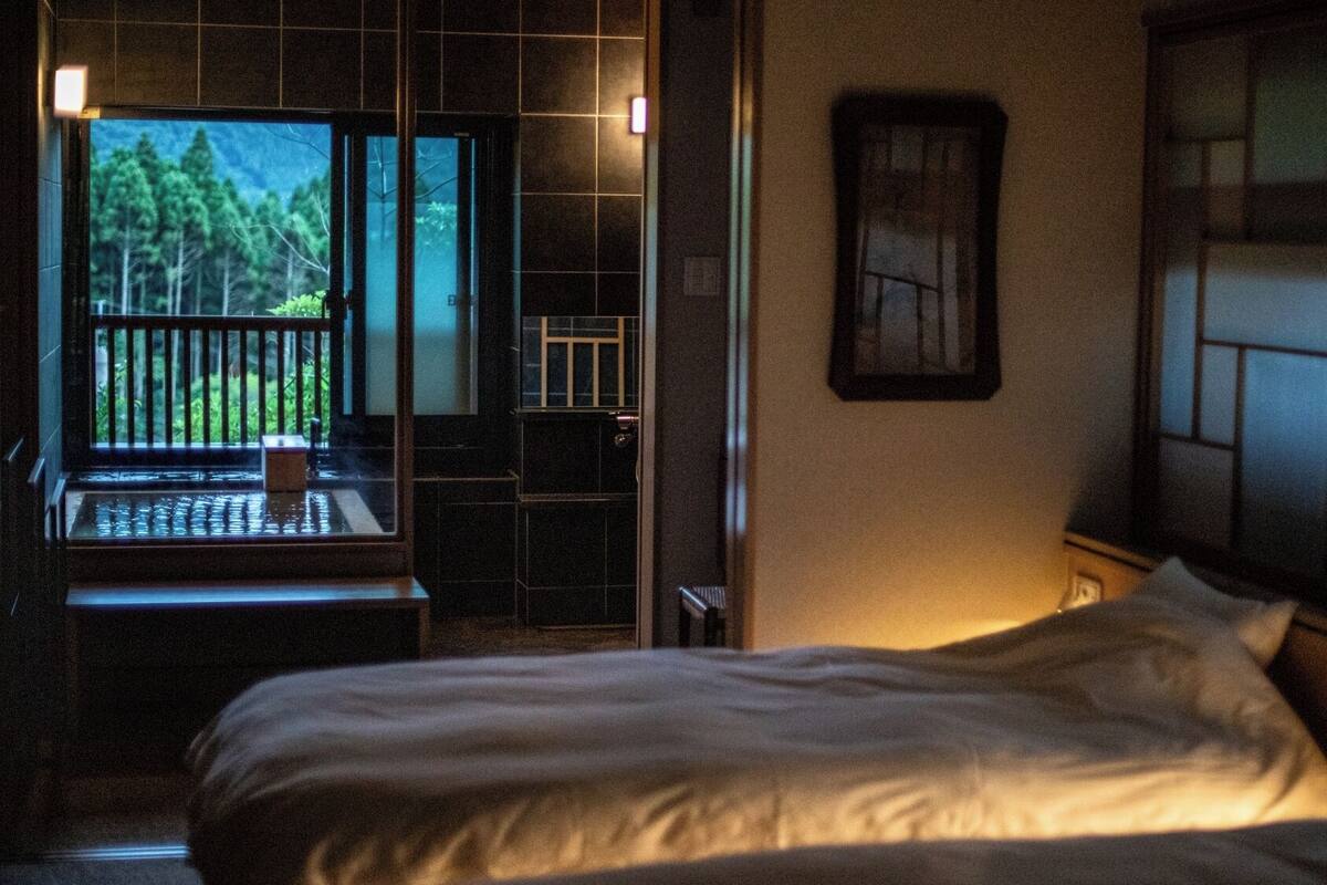 Un dormitorio con una cama, un ventanal con vistas a árboles y una mesita.