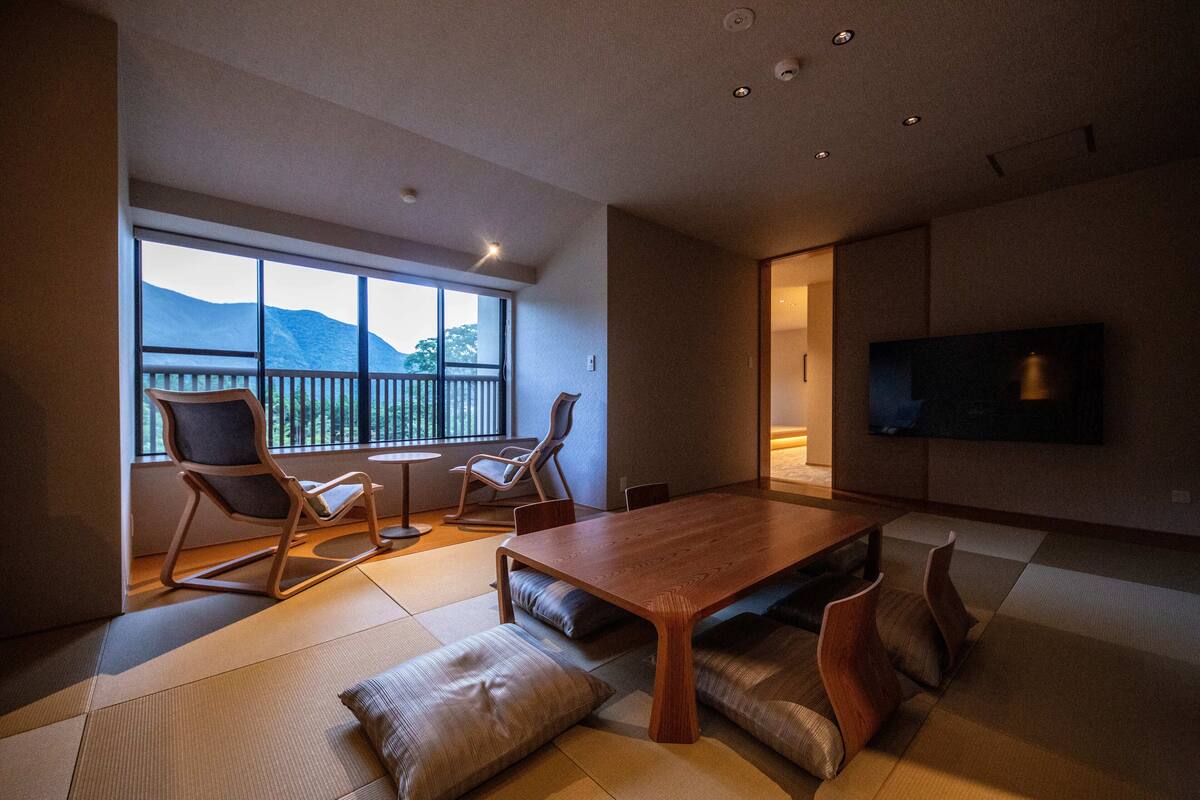 Una sala de estar moderna con una mesa de comedor de madera, asientos acolchados, un televisor y un ventanal con vistas a la montaña.