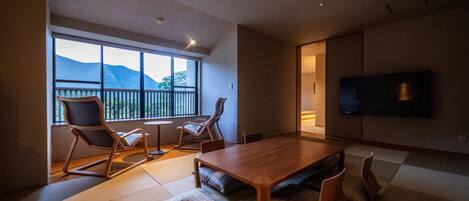 A modern living room with a wooden dining table, cushioned seating, a television, and a large window overlooking a mountain.