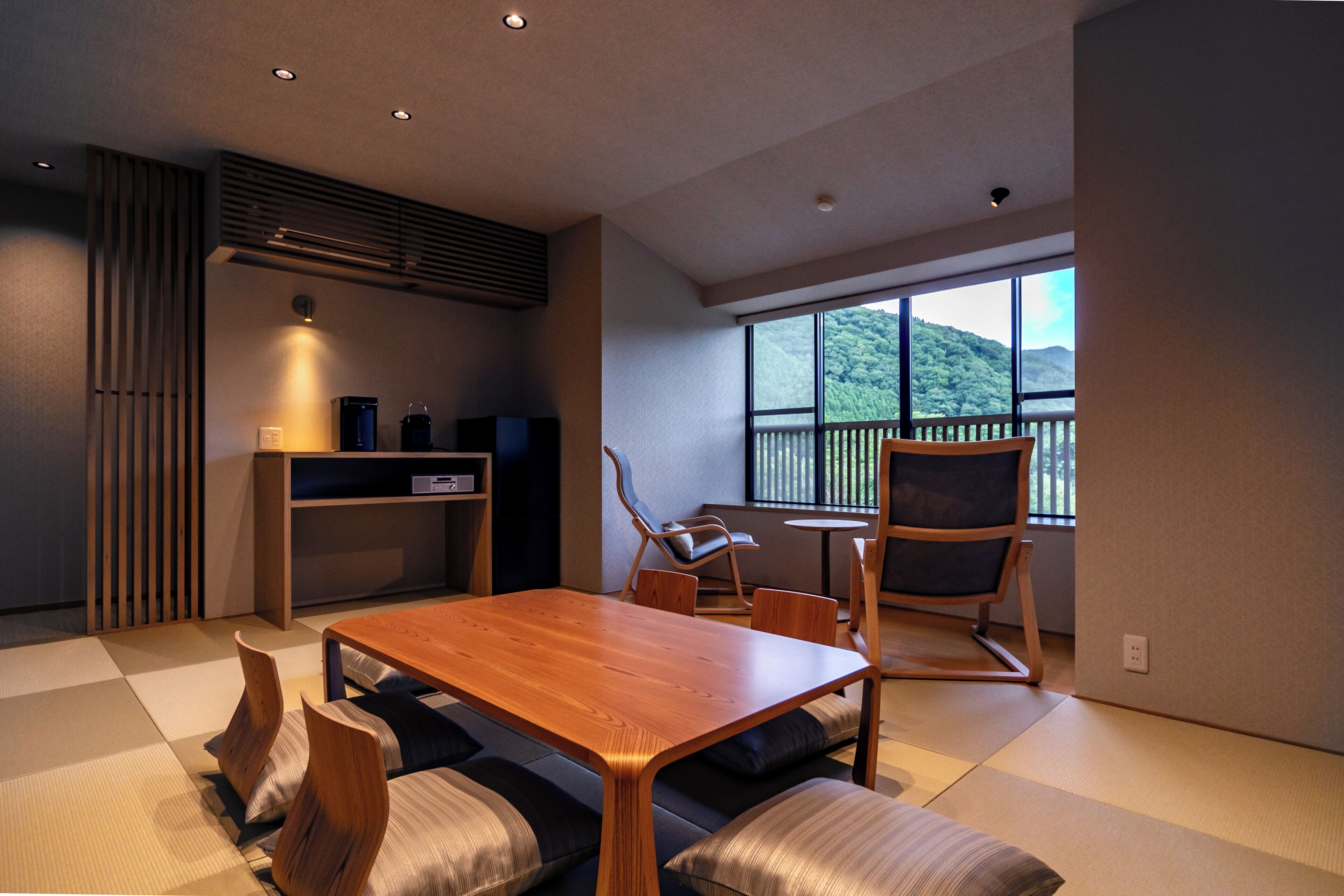A modern room with a wooden dining table, cushioned seating, a small kitchen area, and a large window offering a view of greenery.