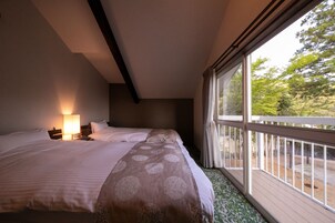 Un dormitorio con una cama grande, una mesita de noche con lámpara y vistas a un balcón con árboles.