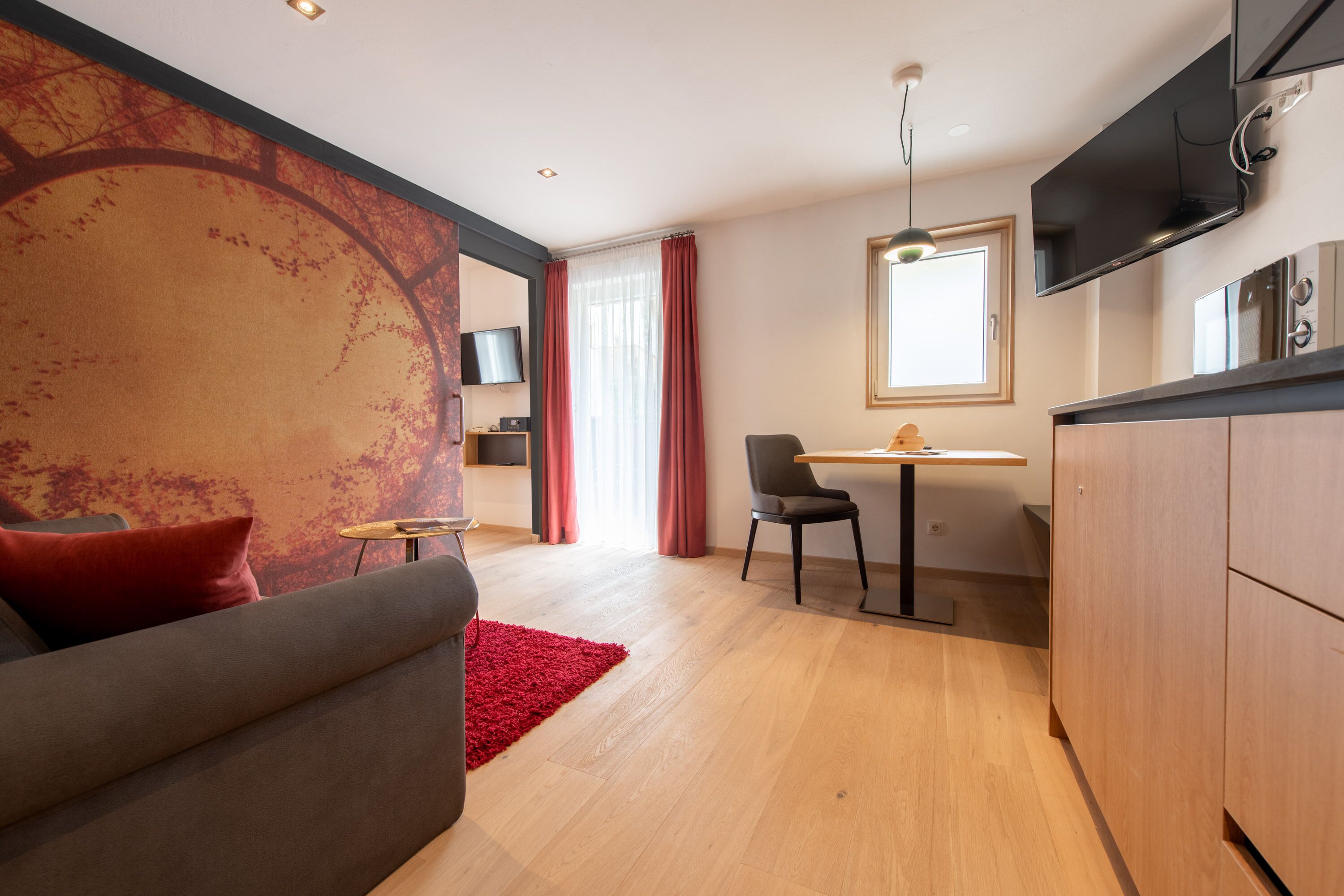 A modern living room with a sofa, a red rug, a dining area with a table and chairs, and a kitchen with a TV mounted above the counter.