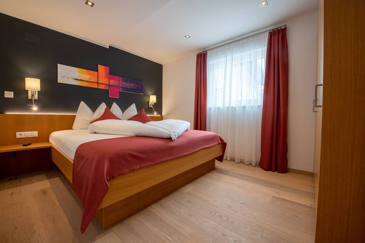 A modern hotel room with a large bed, wooden headboard, red curtains, and a wall-mounted abstract art piece.