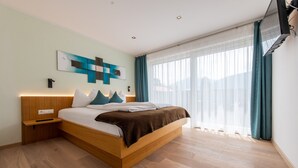 A modern bedroom with a wooden bed, a nightstand, a wall-mounted TV, and a large window with curtains. - Lapis Monti - Apartments & Suites  (Malles Venosta)