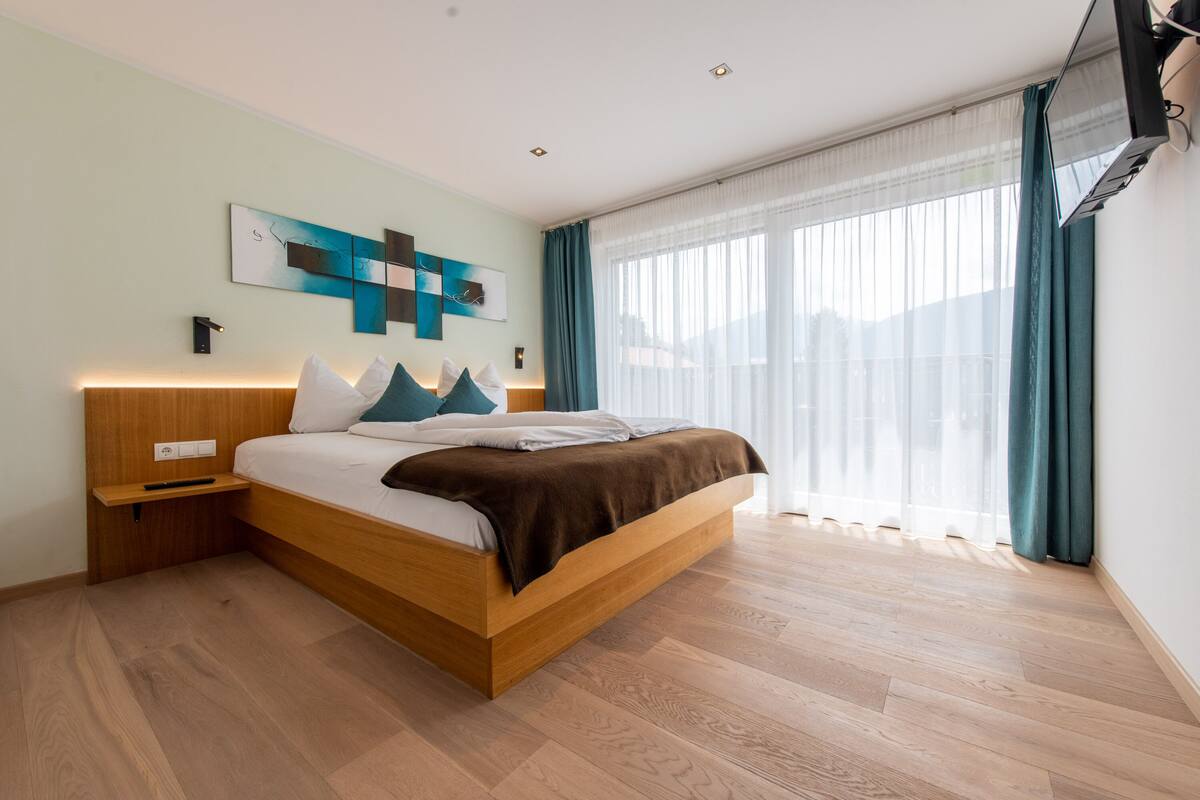 A modern bedroom with a wooden bed, a nightstand, a wall-mounted TV, and a large window with curtains.