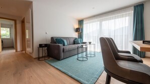 A modern living room with a sofa, two side tables, a chair, and a large window with curtains. - Lapis Monti - Apartments & Suites  (Malles Venosta)