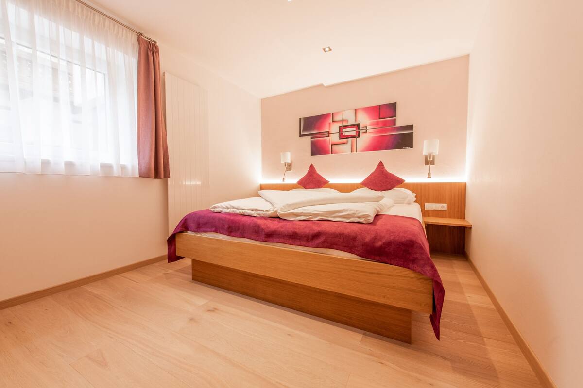 A modern bedroom with a wooden bed, a red blanket, and a wall art piece.