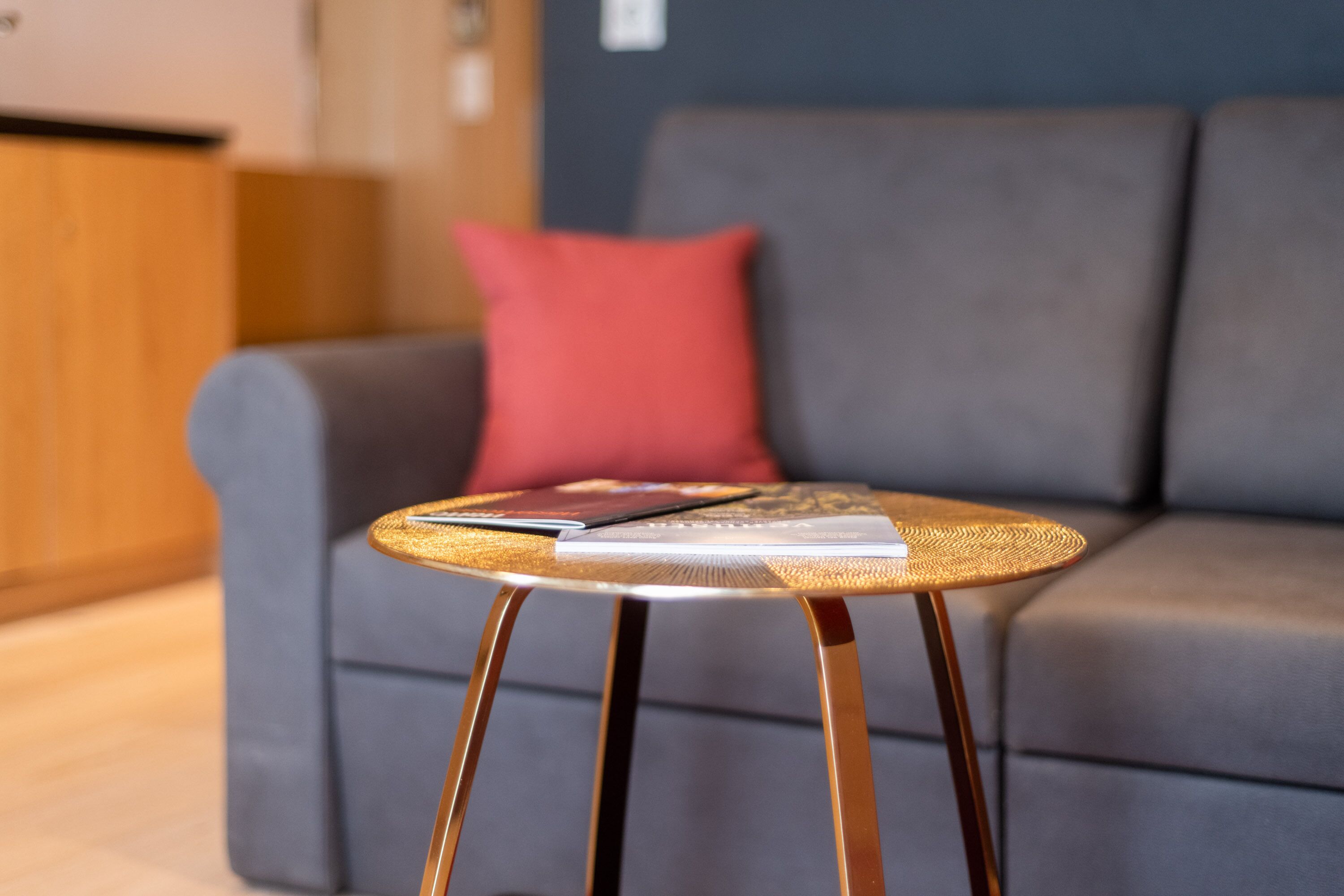 Ein runder, gewebter Couchtisch mit einem Buch darauf, vor einer grauen Couch mit einem roten Kissen platziert.